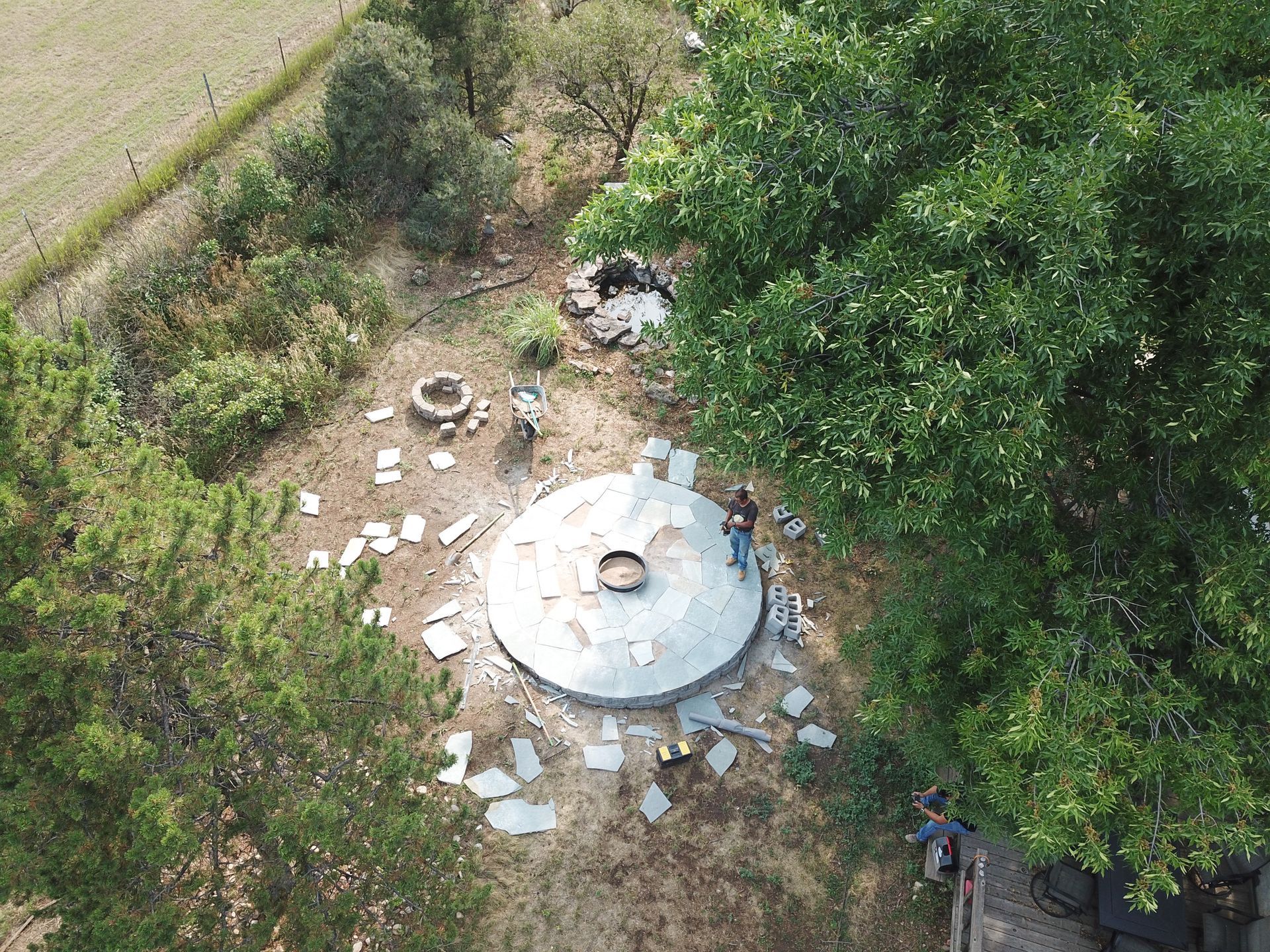 Overhead view of a person standing near a round, metal structure in a wooded area with scattered debris.