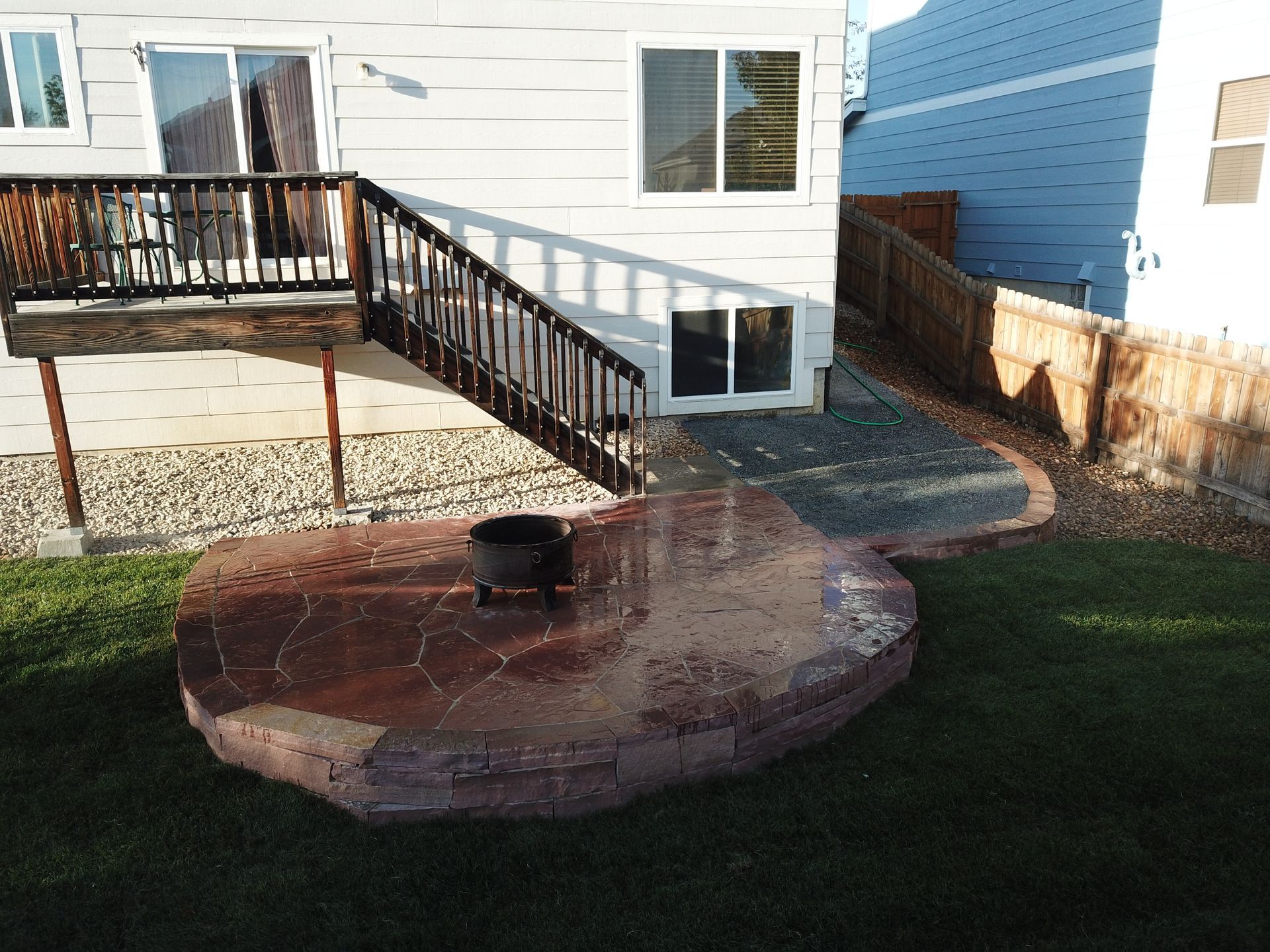 Backyard patio with fire pit, deck, and steps leading down to lawn and landscaping.