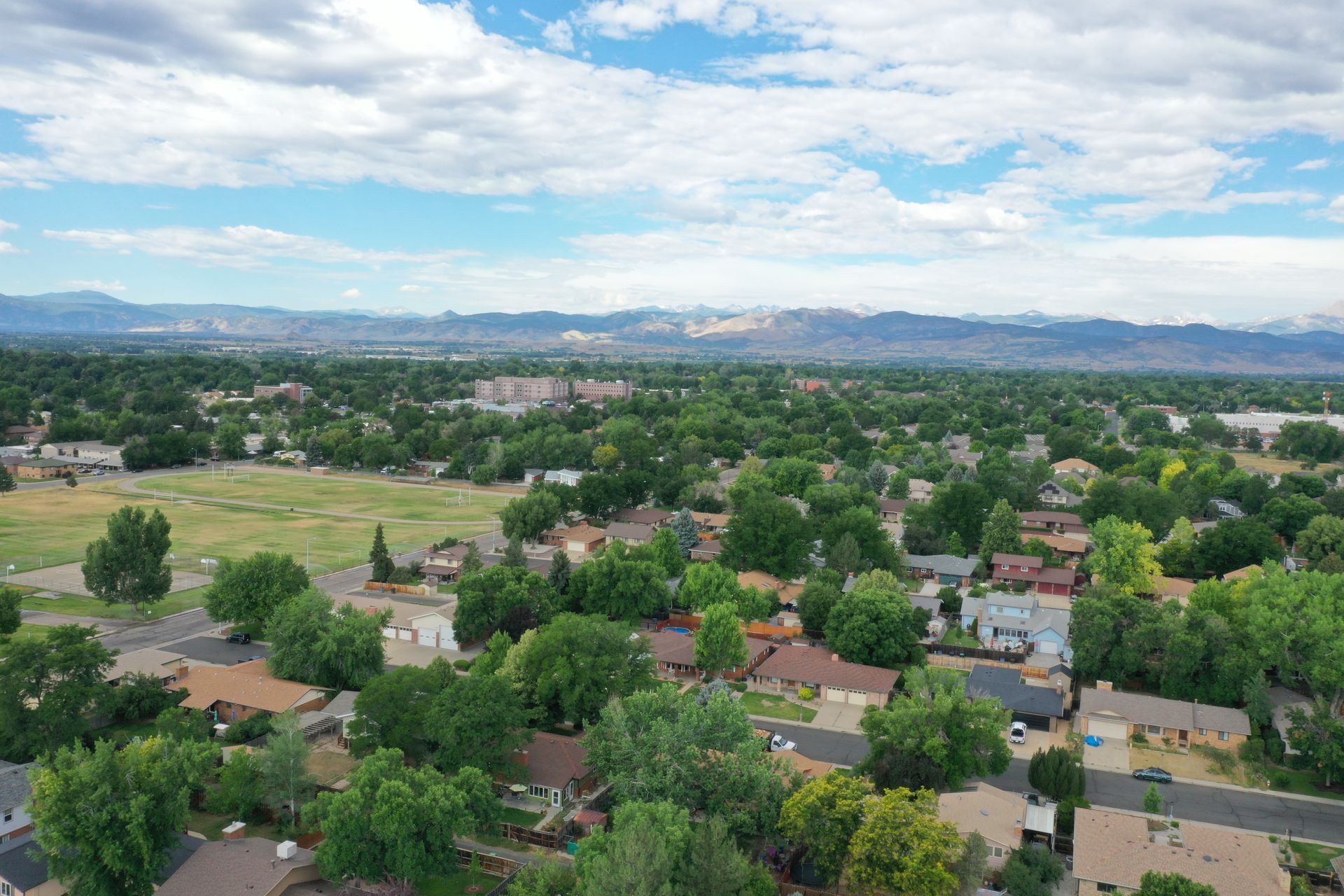 Overhead view of a suburban neighborhood under a partly cloudy blue sky, with mountains in the distance.