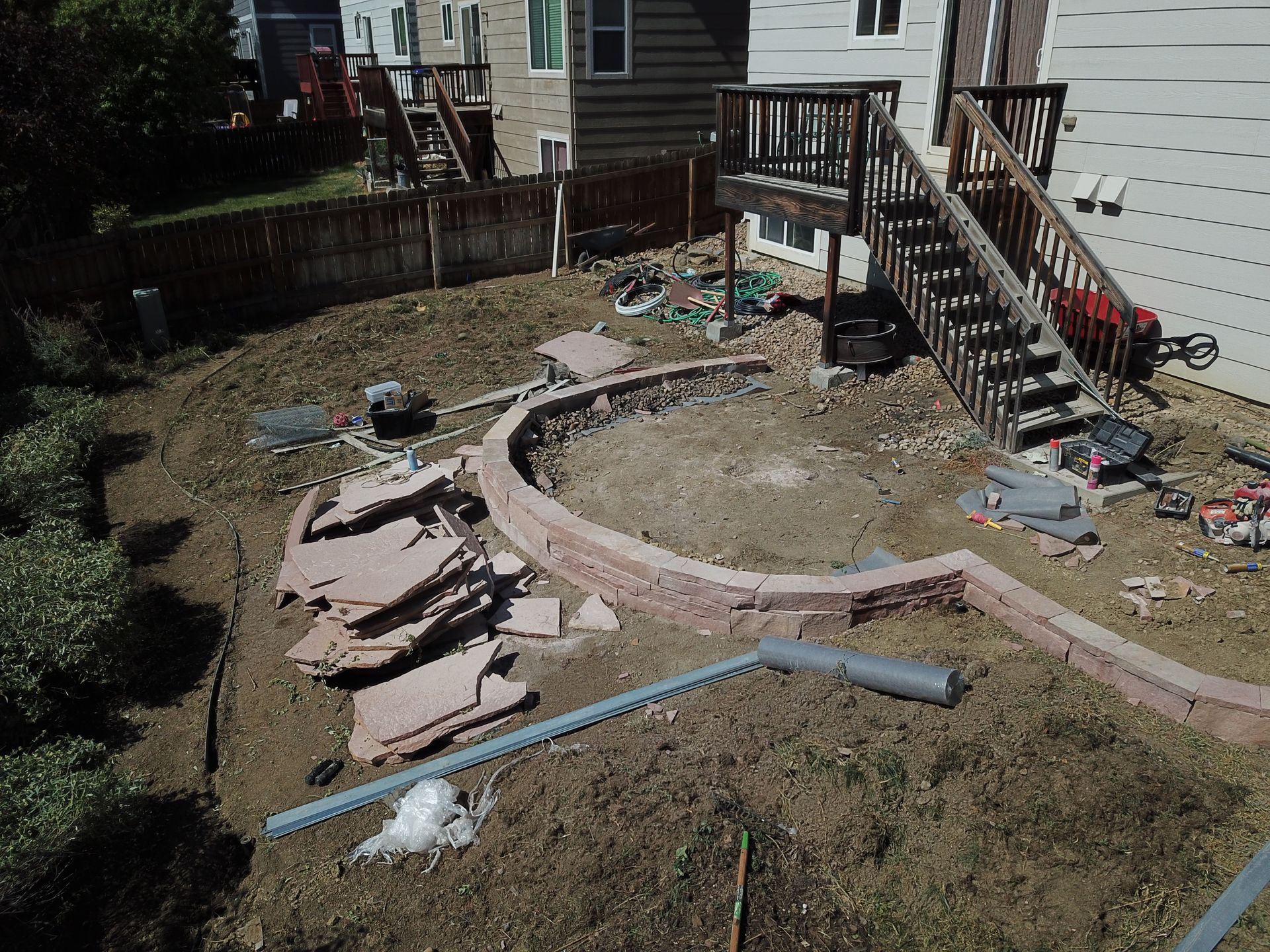 Backyard landscaping project with a circular retaining wall and materials, near a house with a deck and stairs.