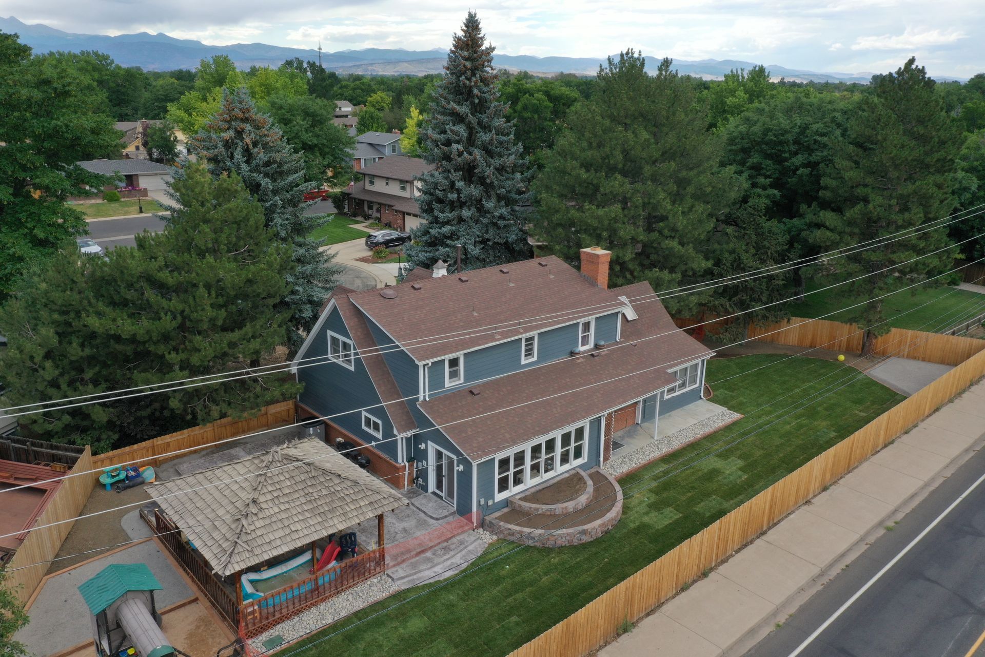 Two-story blue house with brown roof and gazebo in front yard. Surrounded by a wooden fence and trees.