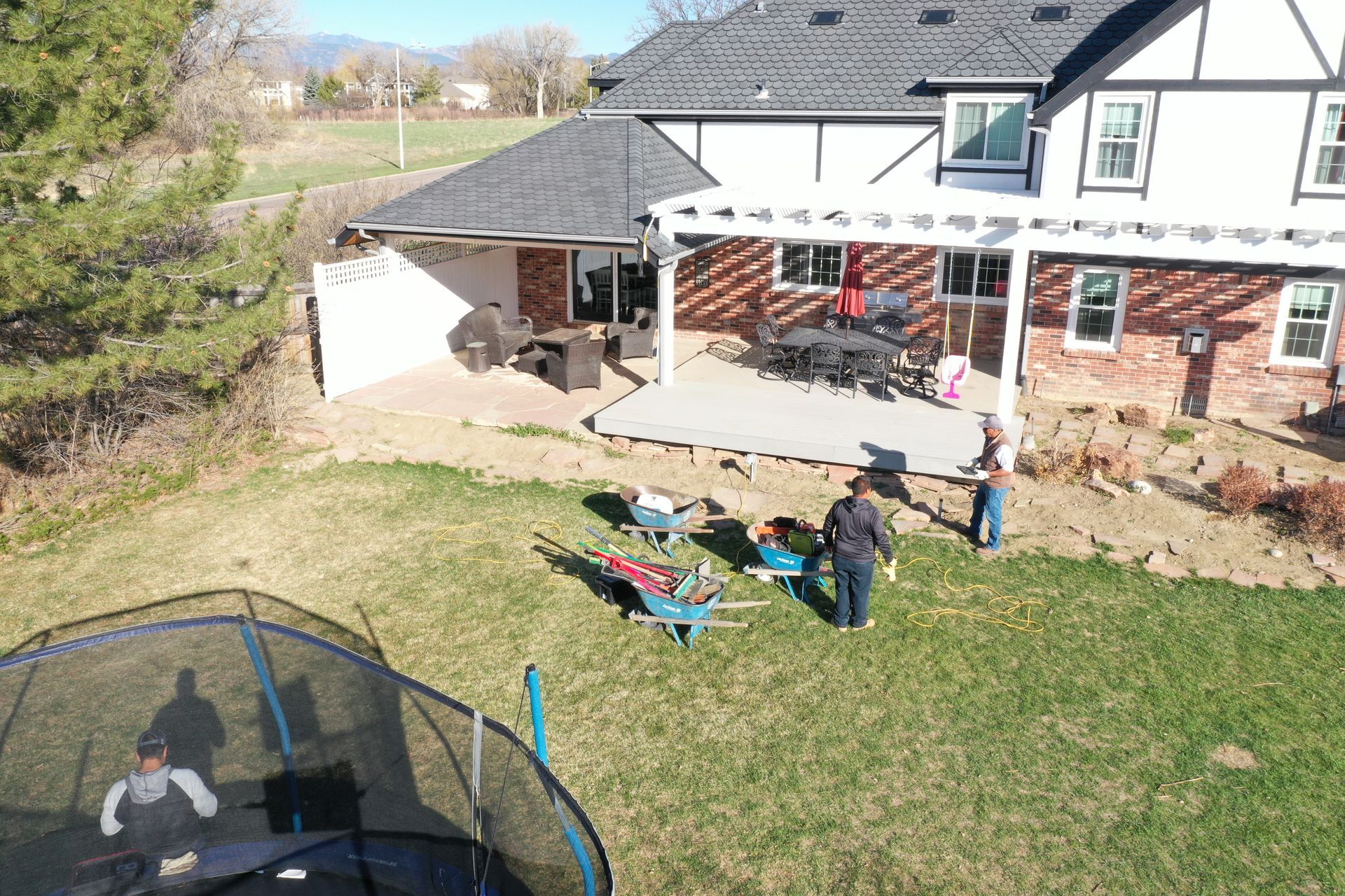 Backyard with a house, patio, and trampoline. People are outdoors, some are jumping on the trampoline.