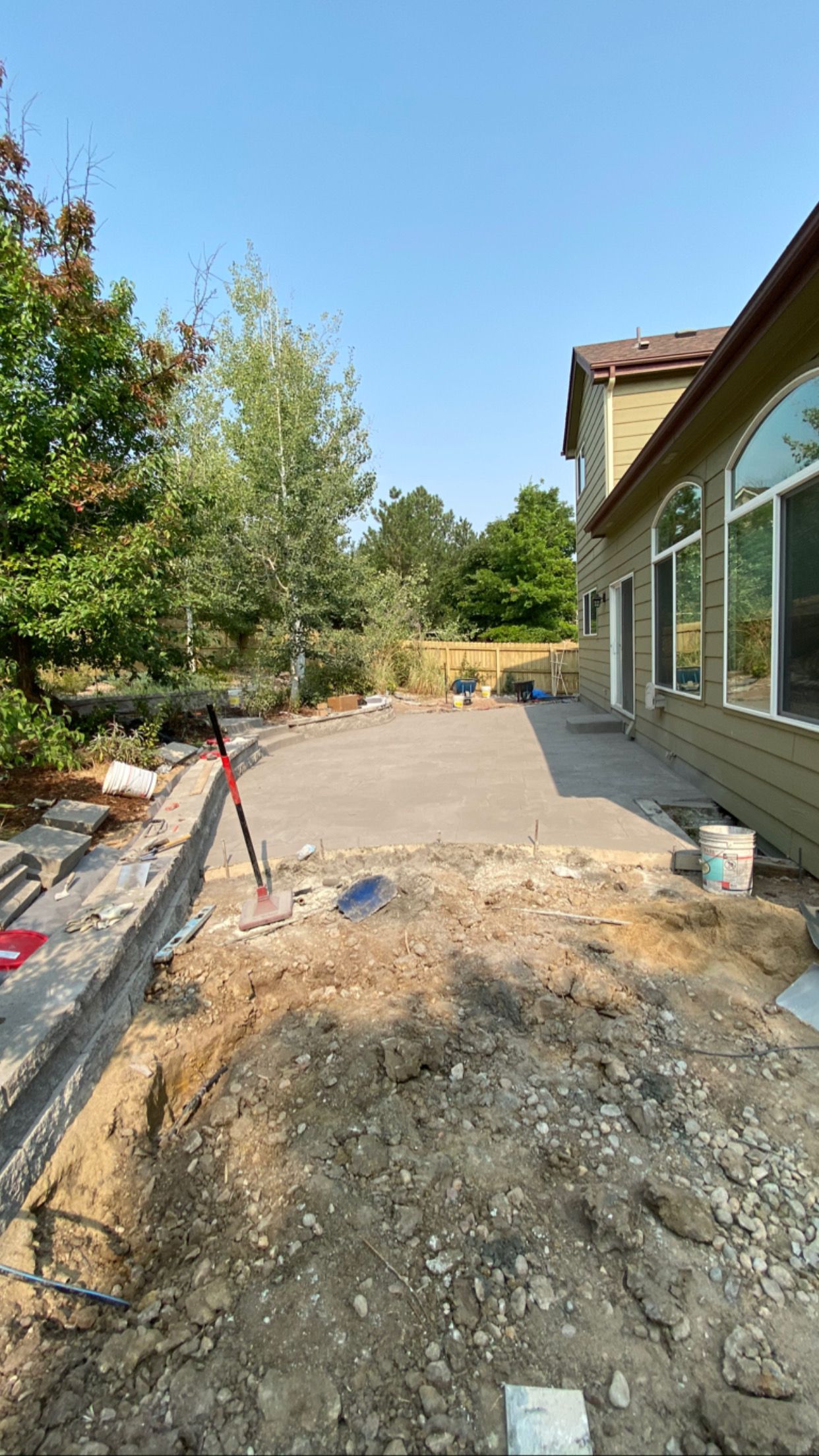 A concrete patio under construction next to a house with exposed dirt and surrounding greenery.