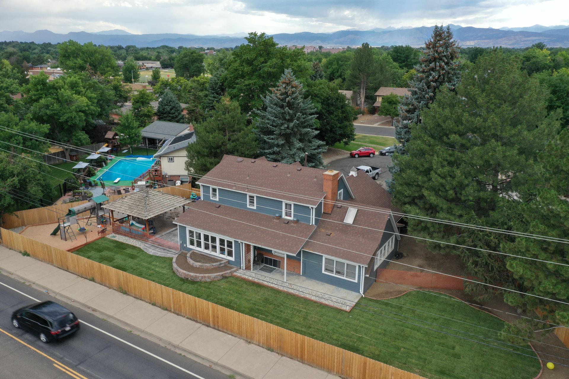 Aerial view of blue house with brown roof and wooden fence, next to a road with a black car.