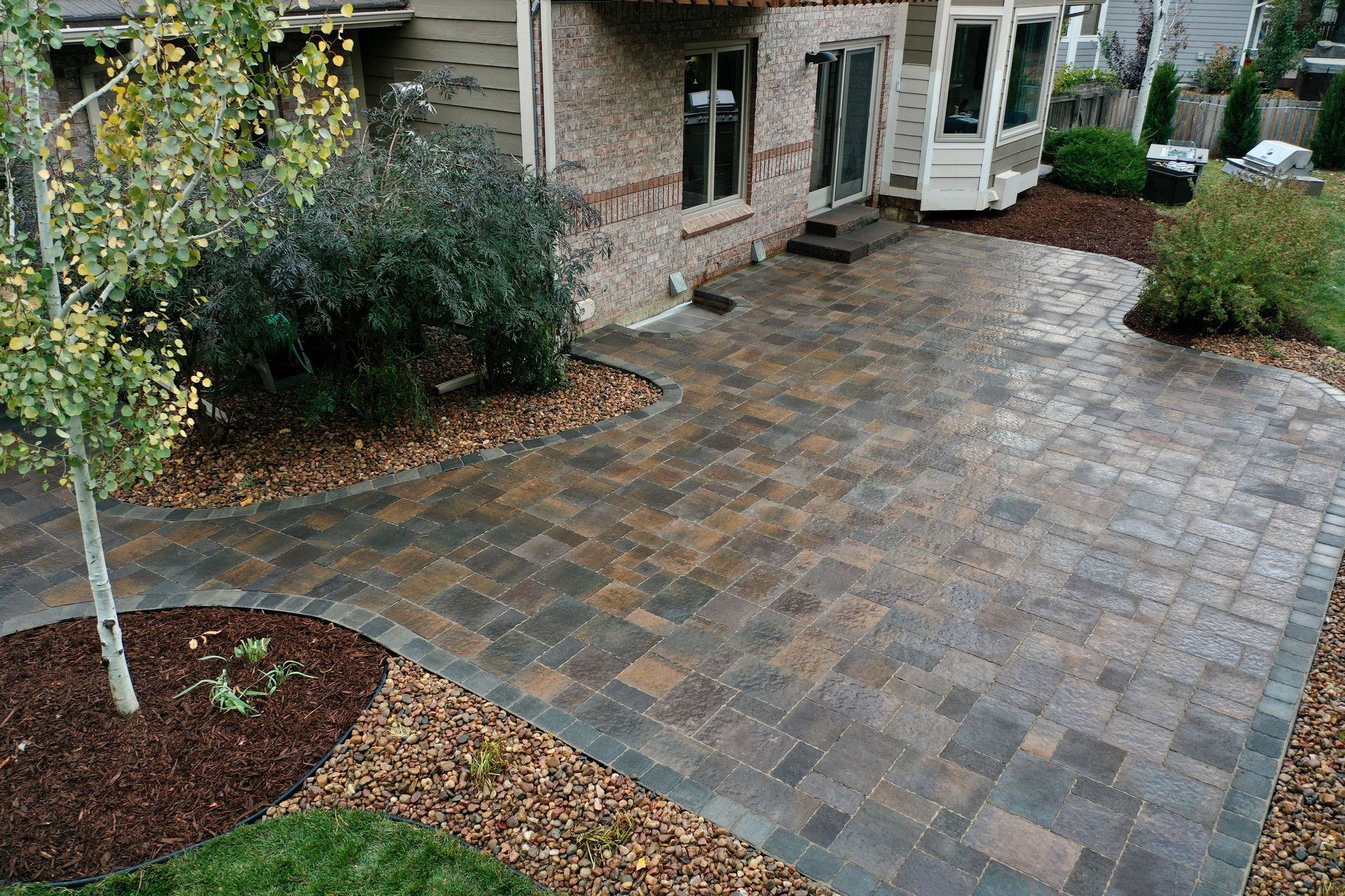 Brick patio with house, garden beds, and a small tree.