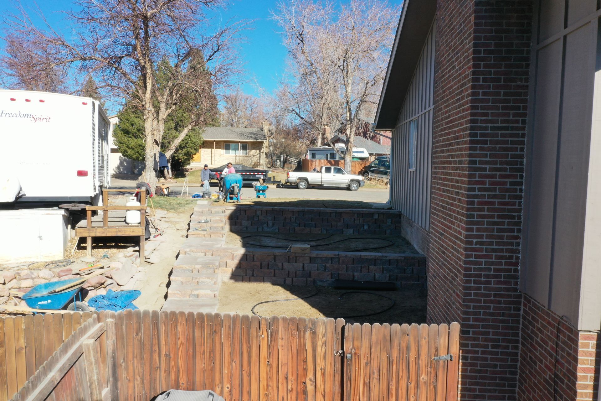 Construction site with retaining wall being built in a residential backyard; people working, truck, camper, fence.
