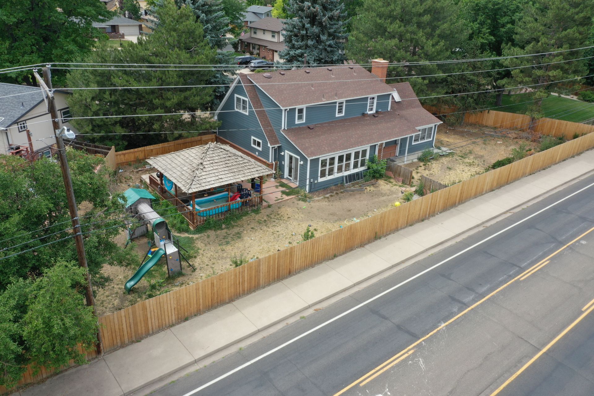 House with blue siding, brown roof, and wooden fence next to a street; a play structure and covered area in the yard.