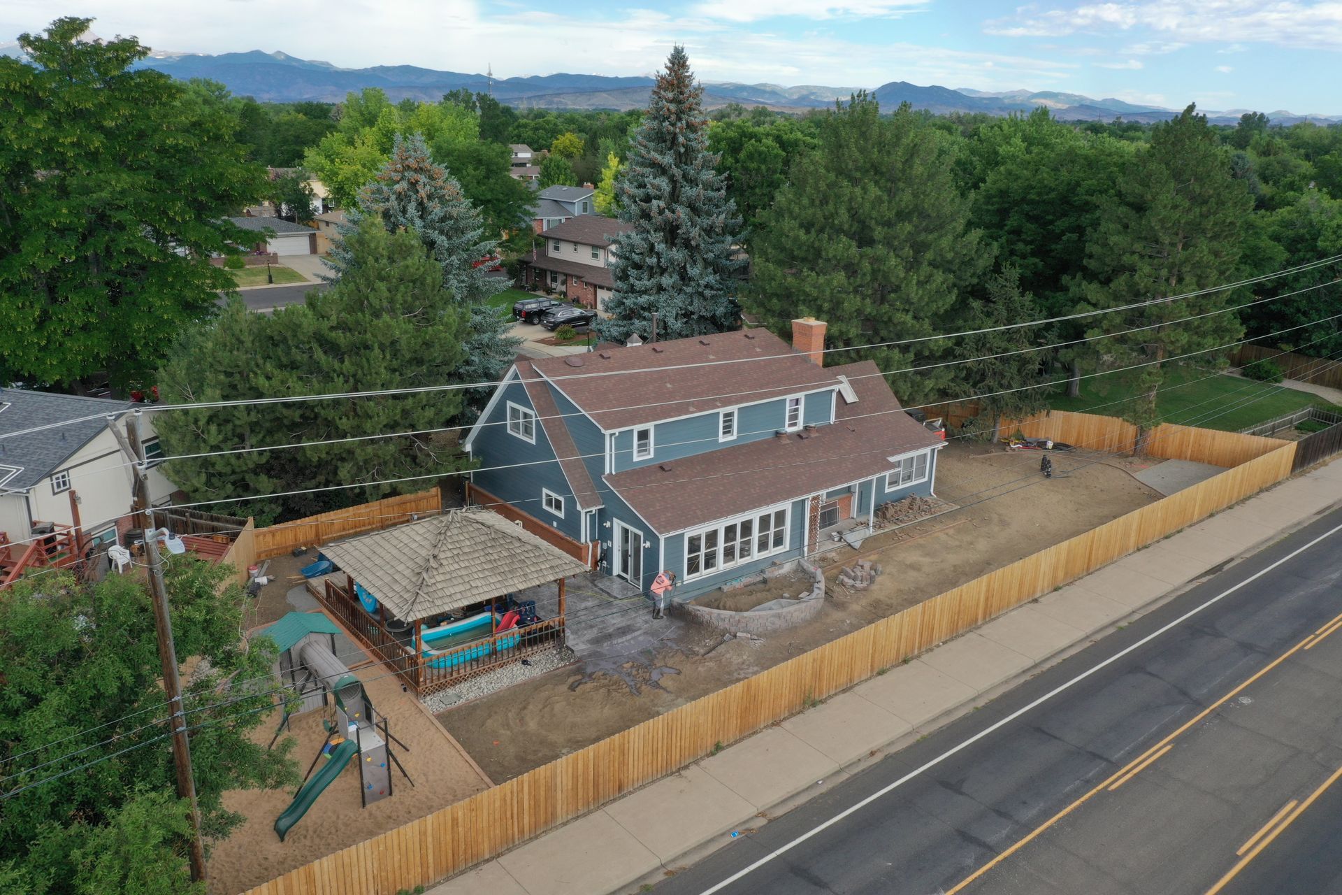 Aerial view of a blue two-story house with a brown roof, wooden fence, and playground next to a road.