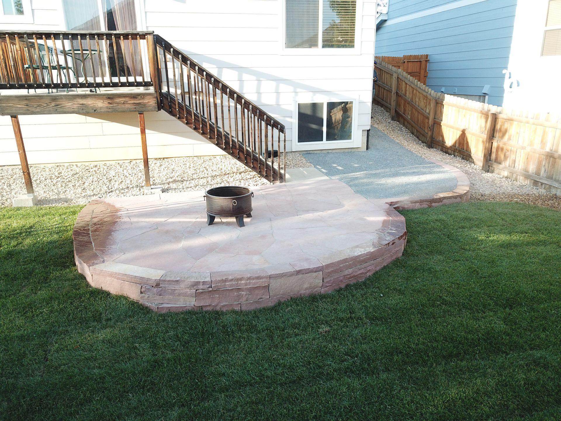 Backyard with a raised, reddish-brown patio with a fire pit, next to a deck and green lawn.