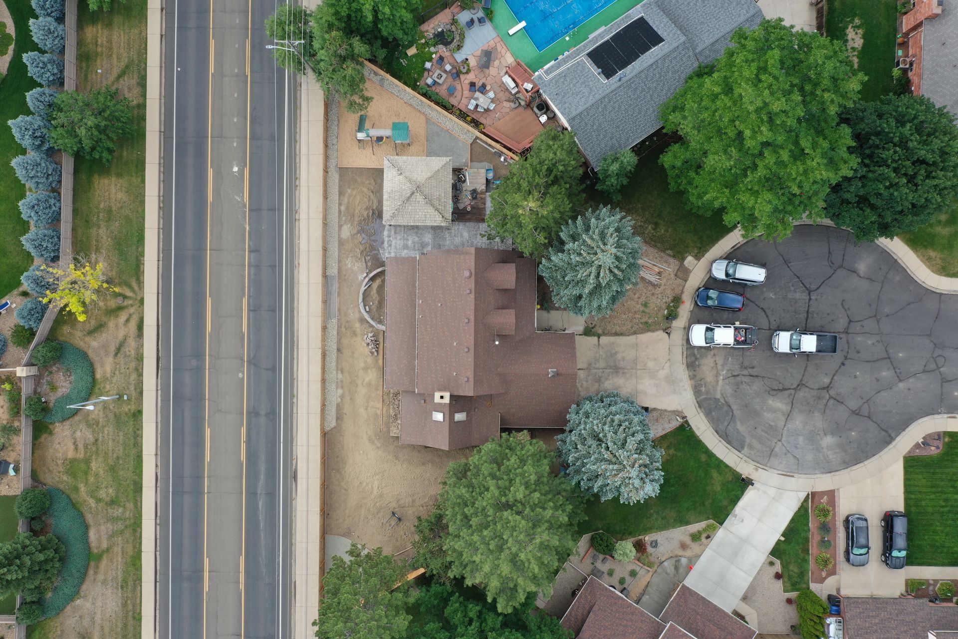 Aerial view of a house next to a road, with a pool, trees, and parked cars visible.