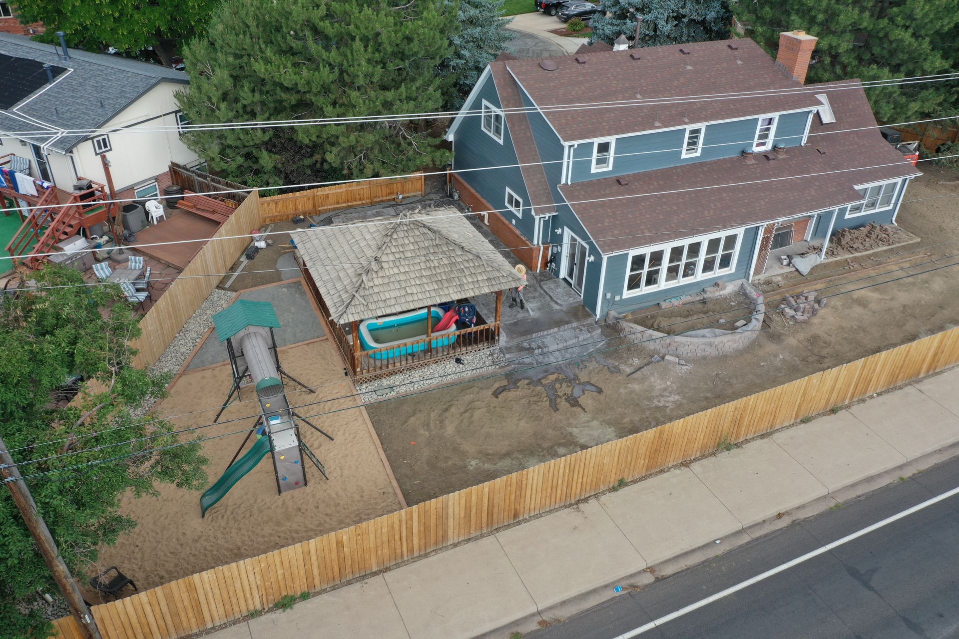 Aerial view of a blue house with a brown roof and a fenced-in backyard with a gazebo, playground, and brown fence.