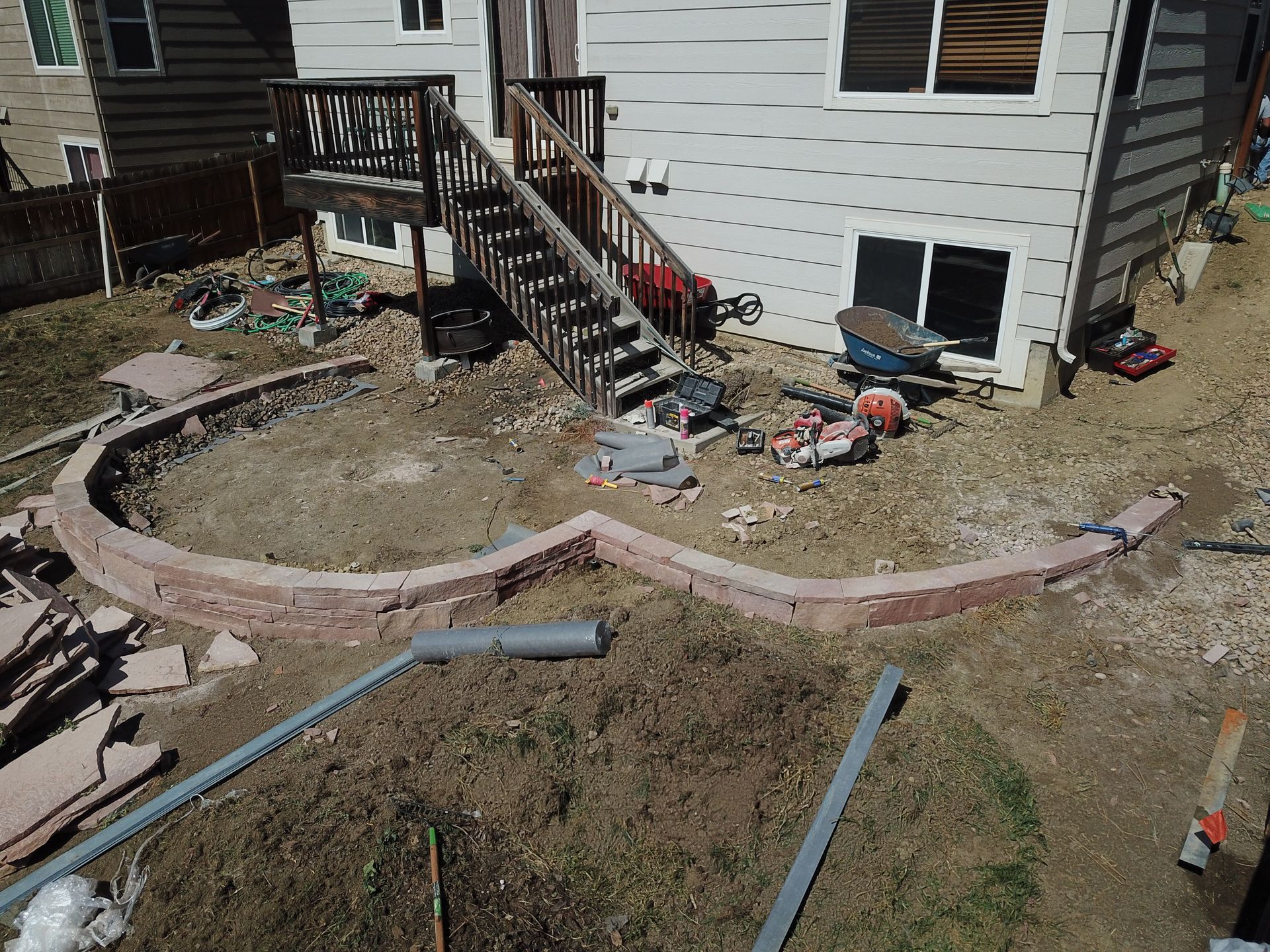 Backyard with a brick retaining wall under construction. Wooden stairs lead to a house. Dirt and tools are visible.