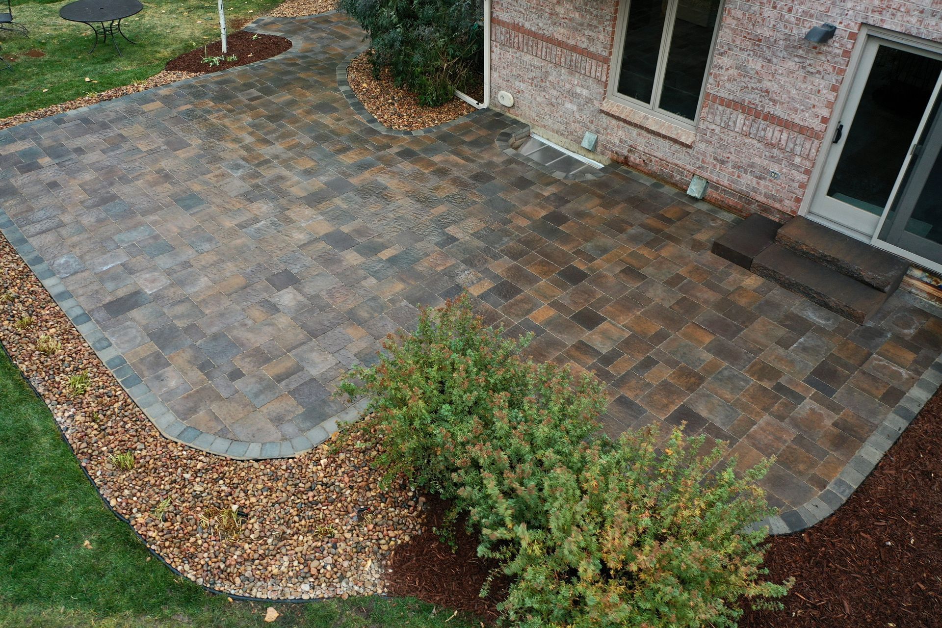 Brick patio adjacent to a brick house, bordered by plants and a bed of brown stones.