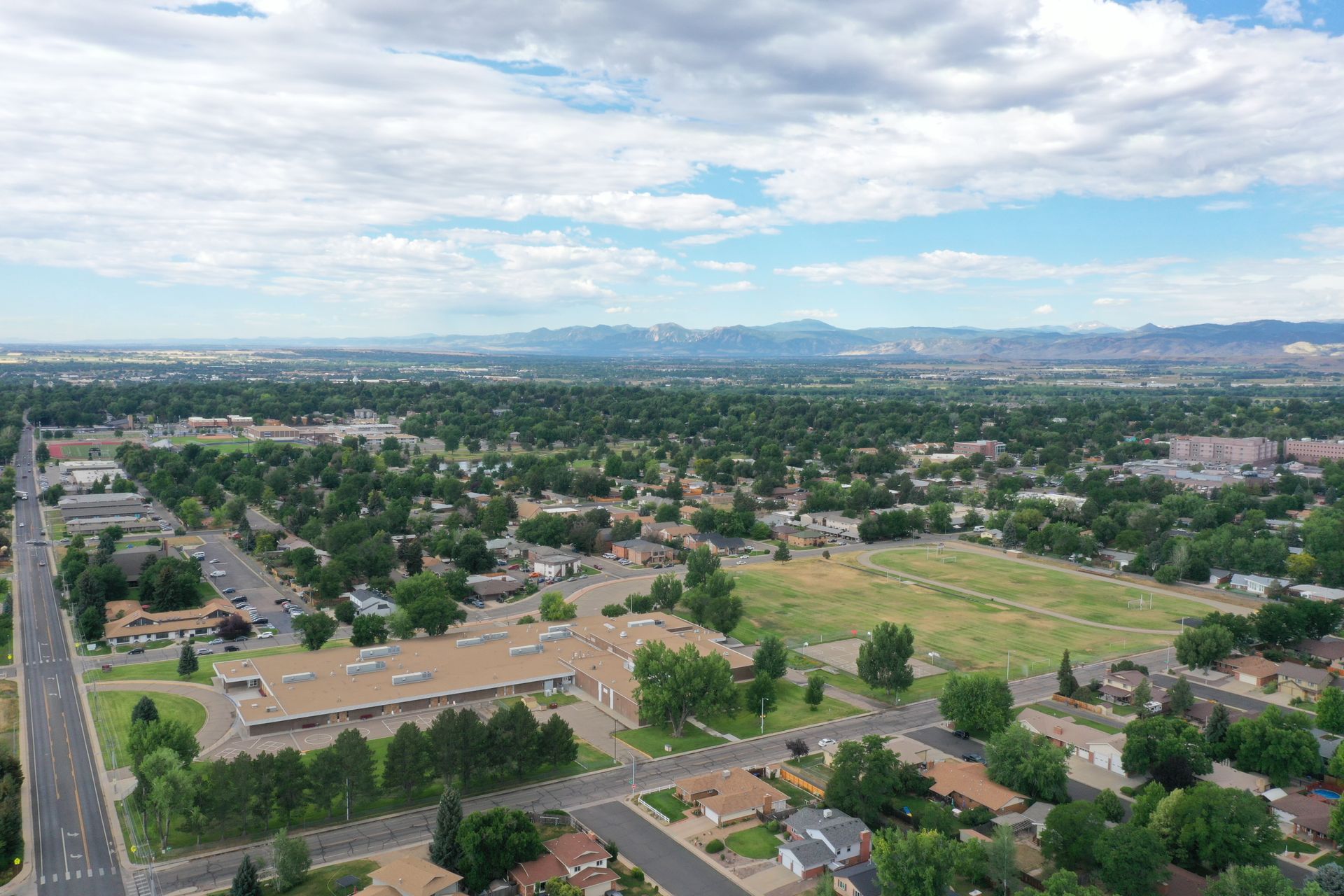Aerial view of a suburban area with green trees, buildings, roads, and a cloudy sky. Mountains visible in the distance.
