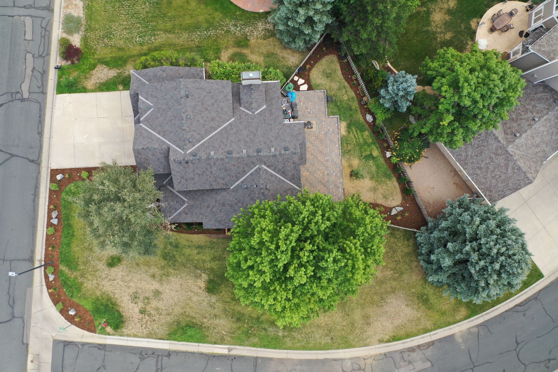 Aerial view of a house with a dark roof and surrounding green trees and grass, on a suburban street.