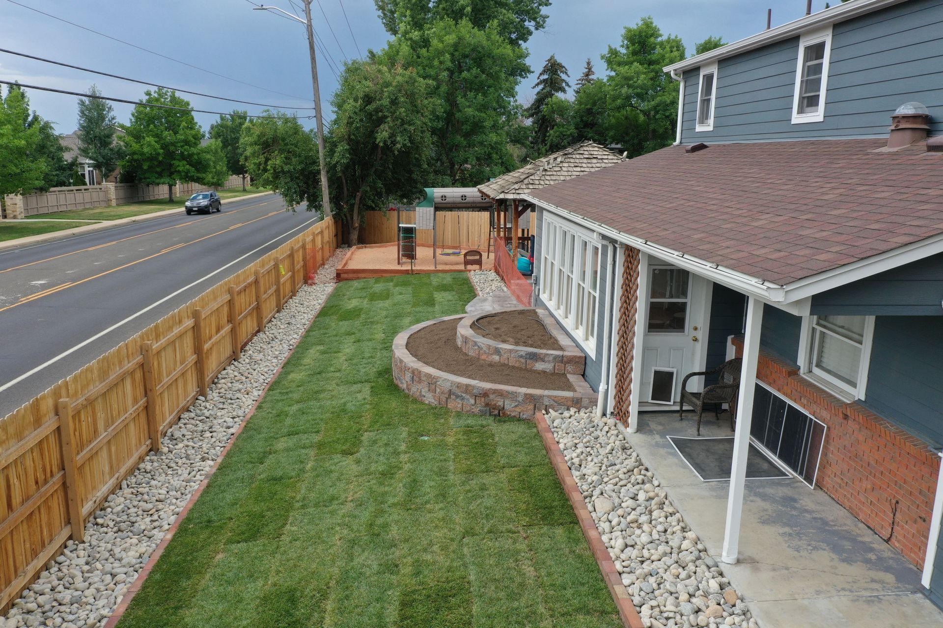 Side view of a house with a green lawn, wooden fence, and paved walkway next to a road.