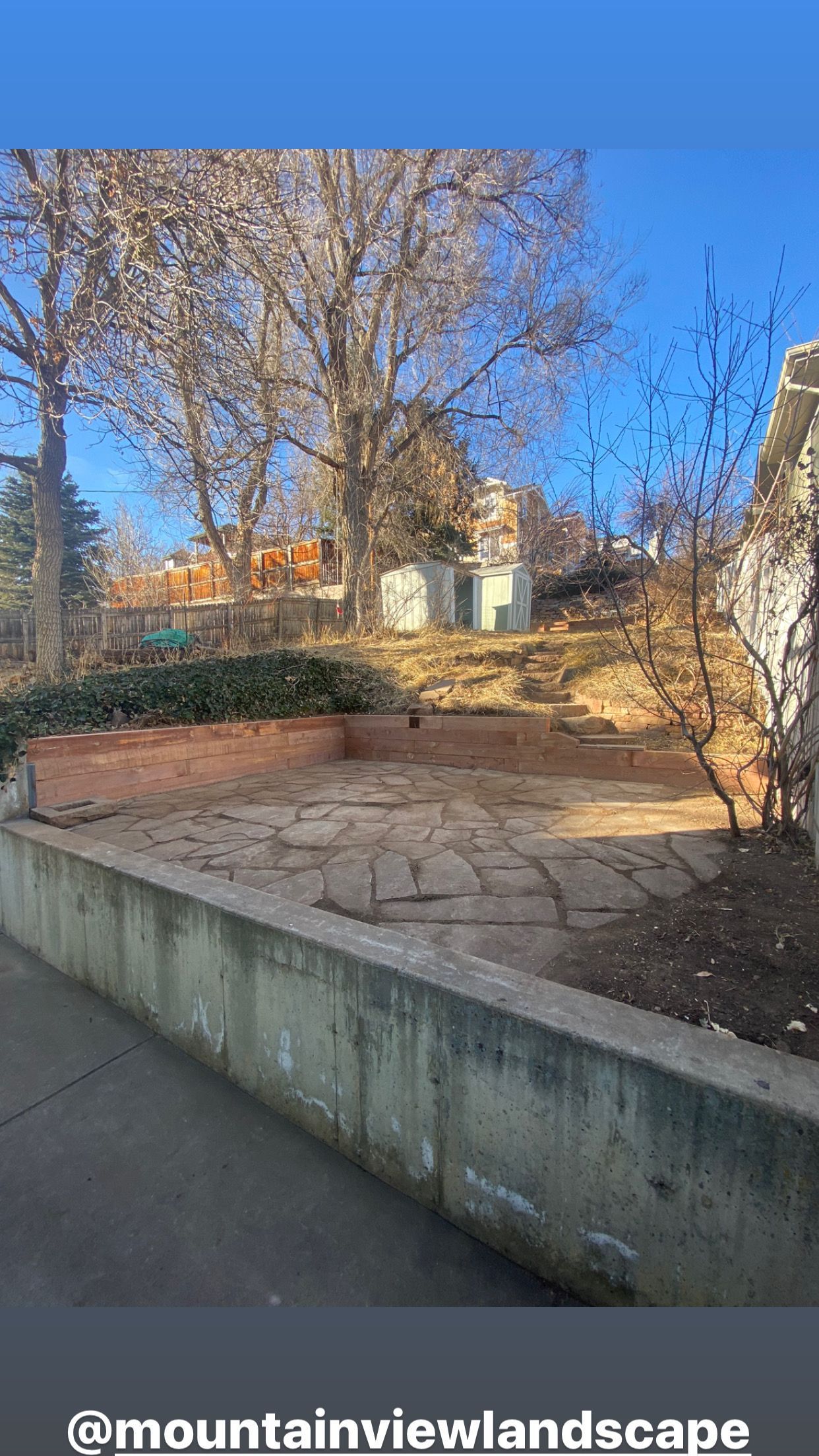 A low concrete wall and paved area, bare trees, and buildings against a blue sky.