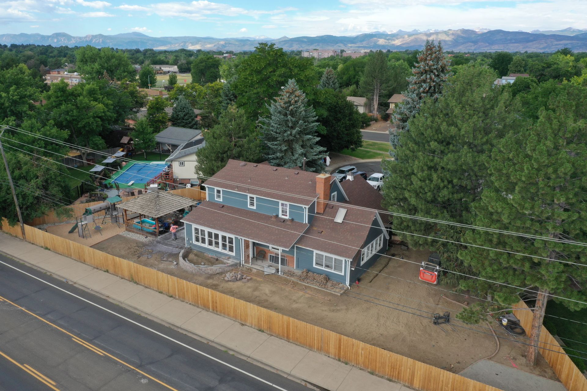 Aerial view of a blue house with a brown roof and a wooden fence along a road, trees and mountains in the background.