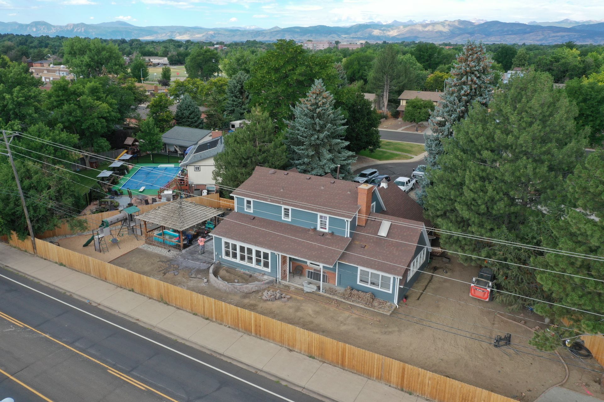 Aerial view of a blue house with a brown roof and a wooden fence, street in front, trees in the background.