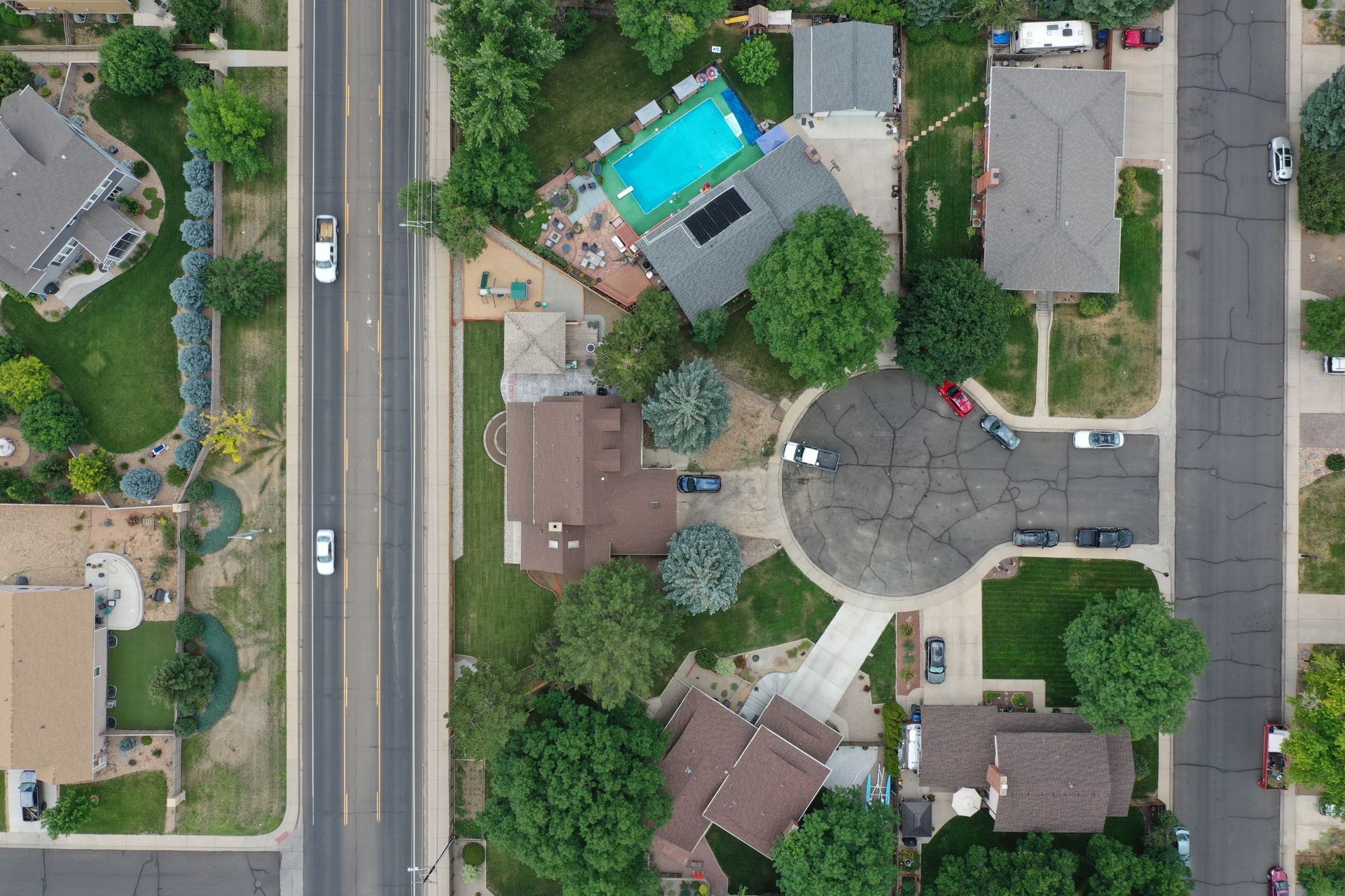 Aerial view of houses and a road. One house has a pool. Green trees and lawns.