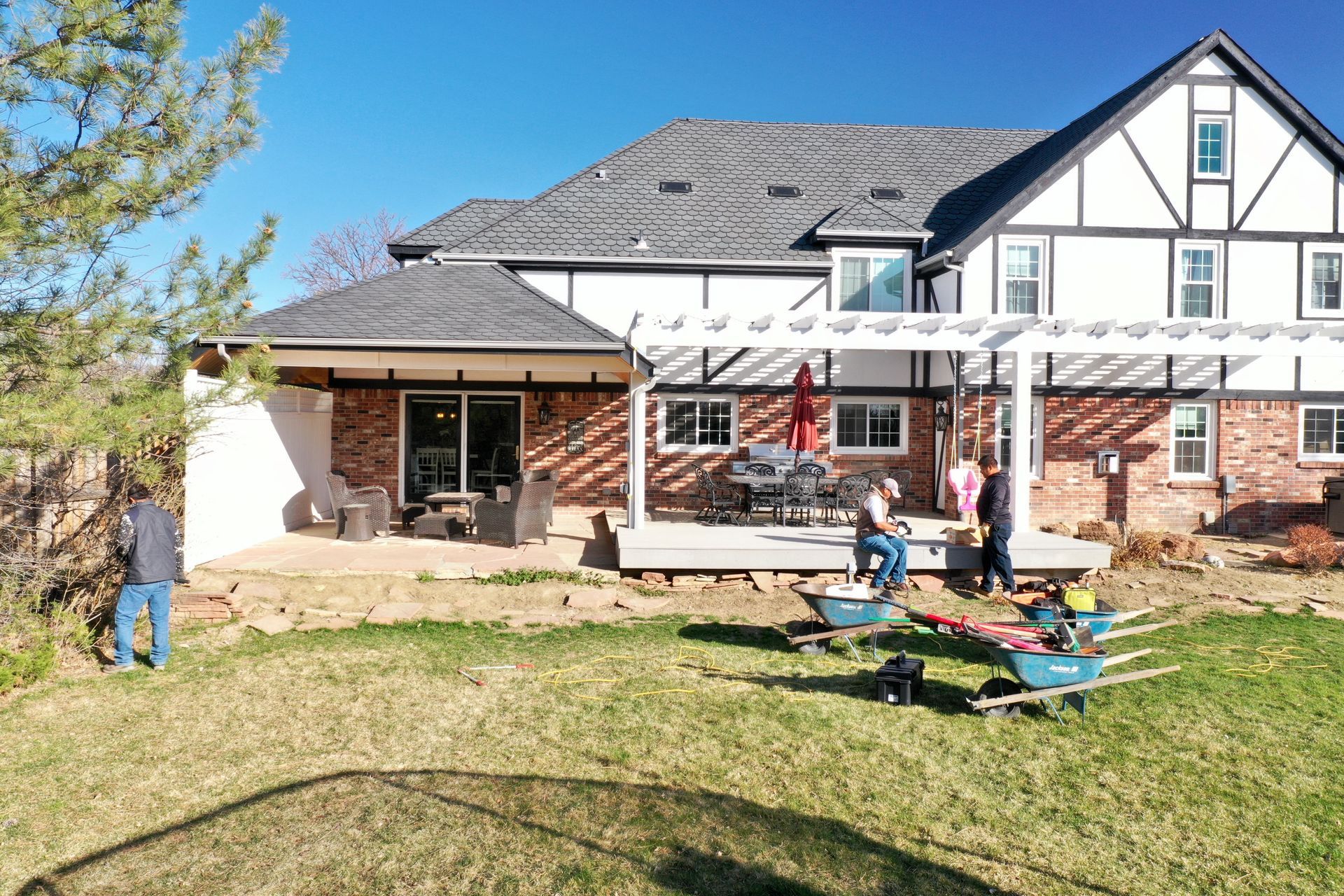 People working on a backyard patio, building a pergola in front of a two-story Tudor house.