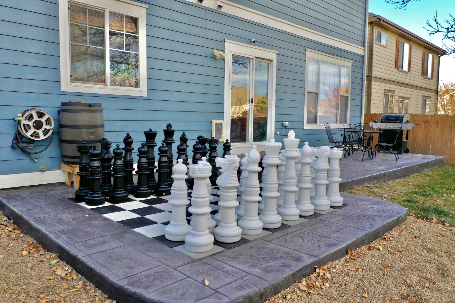 Giant chess set on a patio, black and white pieces, blue house in background.