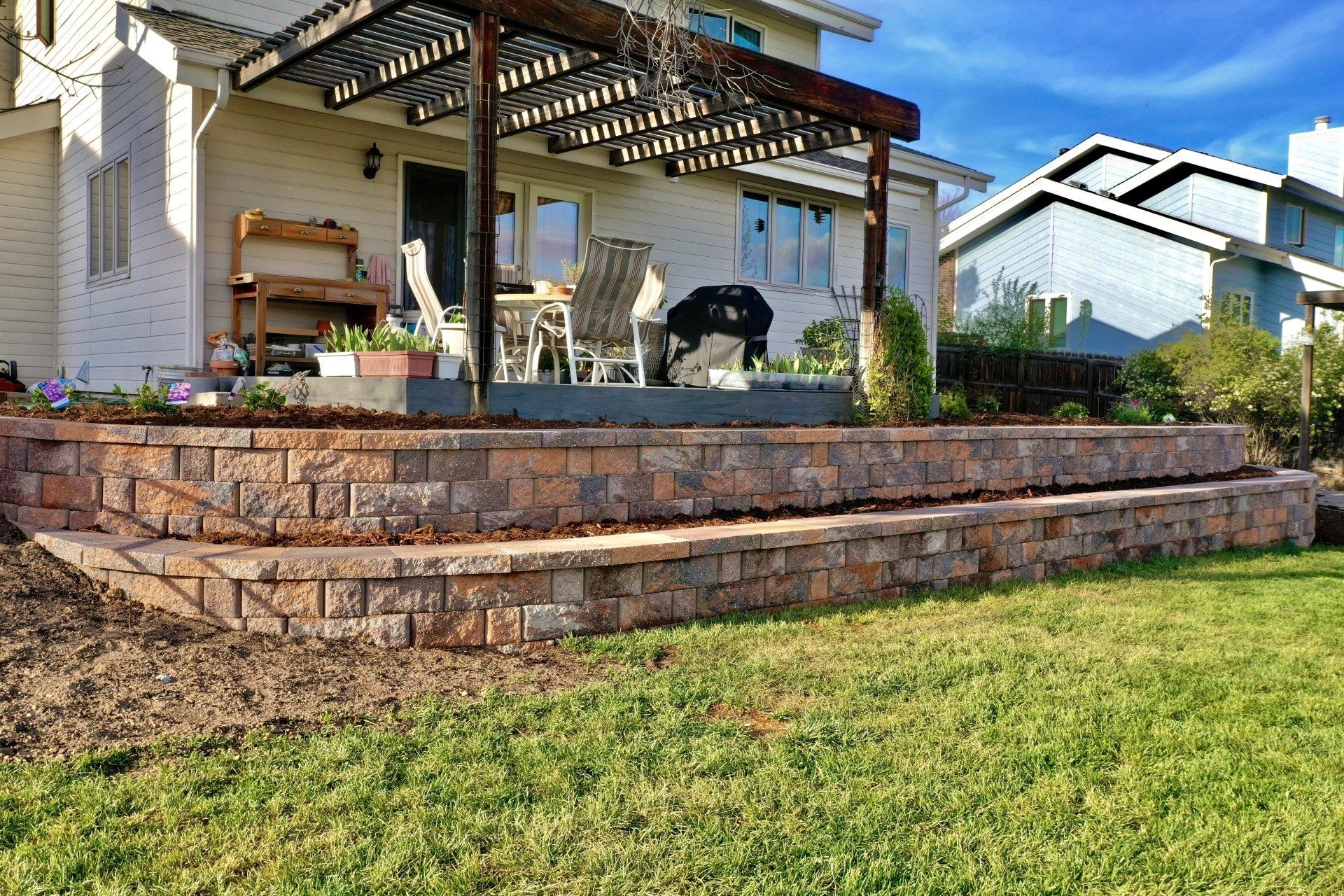 Two-tiered brick retaining wall in front of a house's patio, with pergola and furniture. Green grass and blue sky.