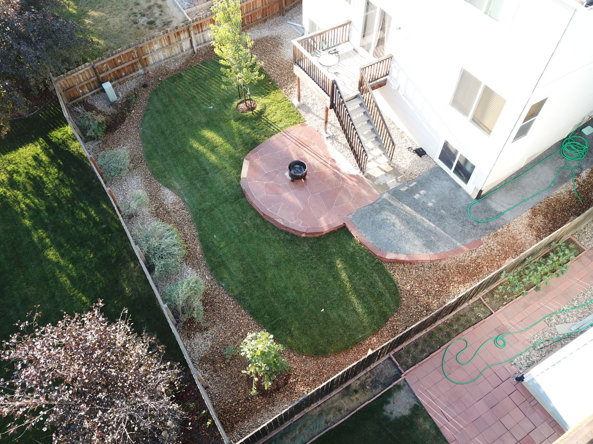 Overhead view of a backyard with a deck, green lawn, fire pit, and landscaping around a two-story house.