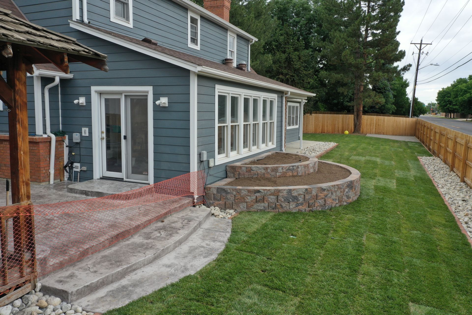 Backyard with teal house, raised stone patio, green lawn, wooden fence, and concrete steps.