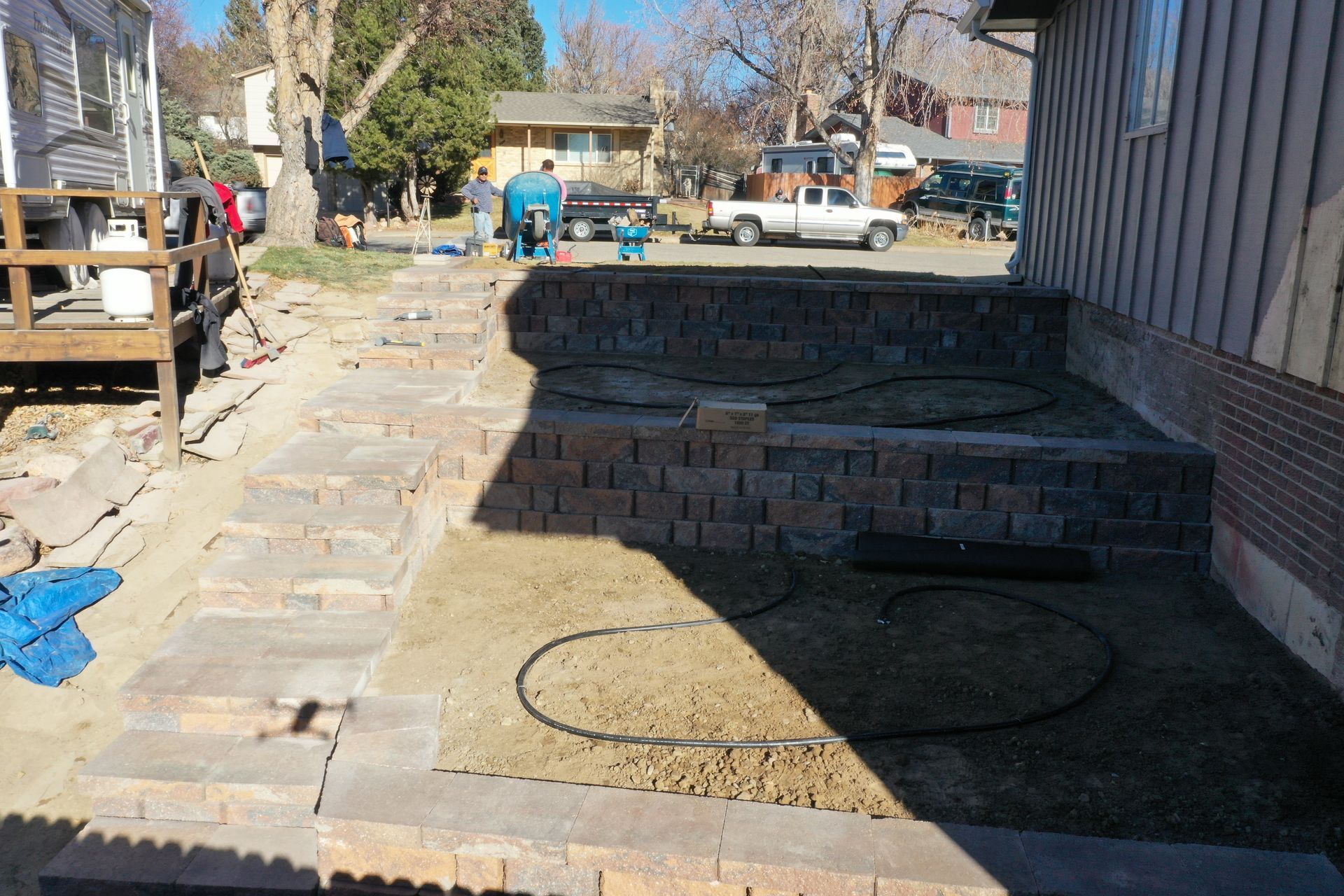 Brick retaining walls and steps in a yard, with a raised garden bed and parked vehicles in the background.