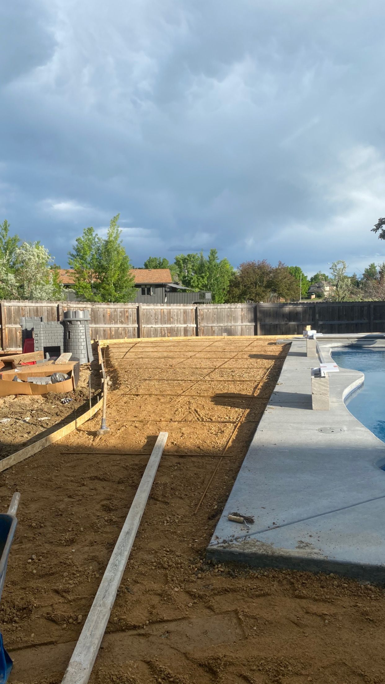 Backyard construction: Dirt area next to a pool, framed by wood and fence; cloudy sky overhead.
