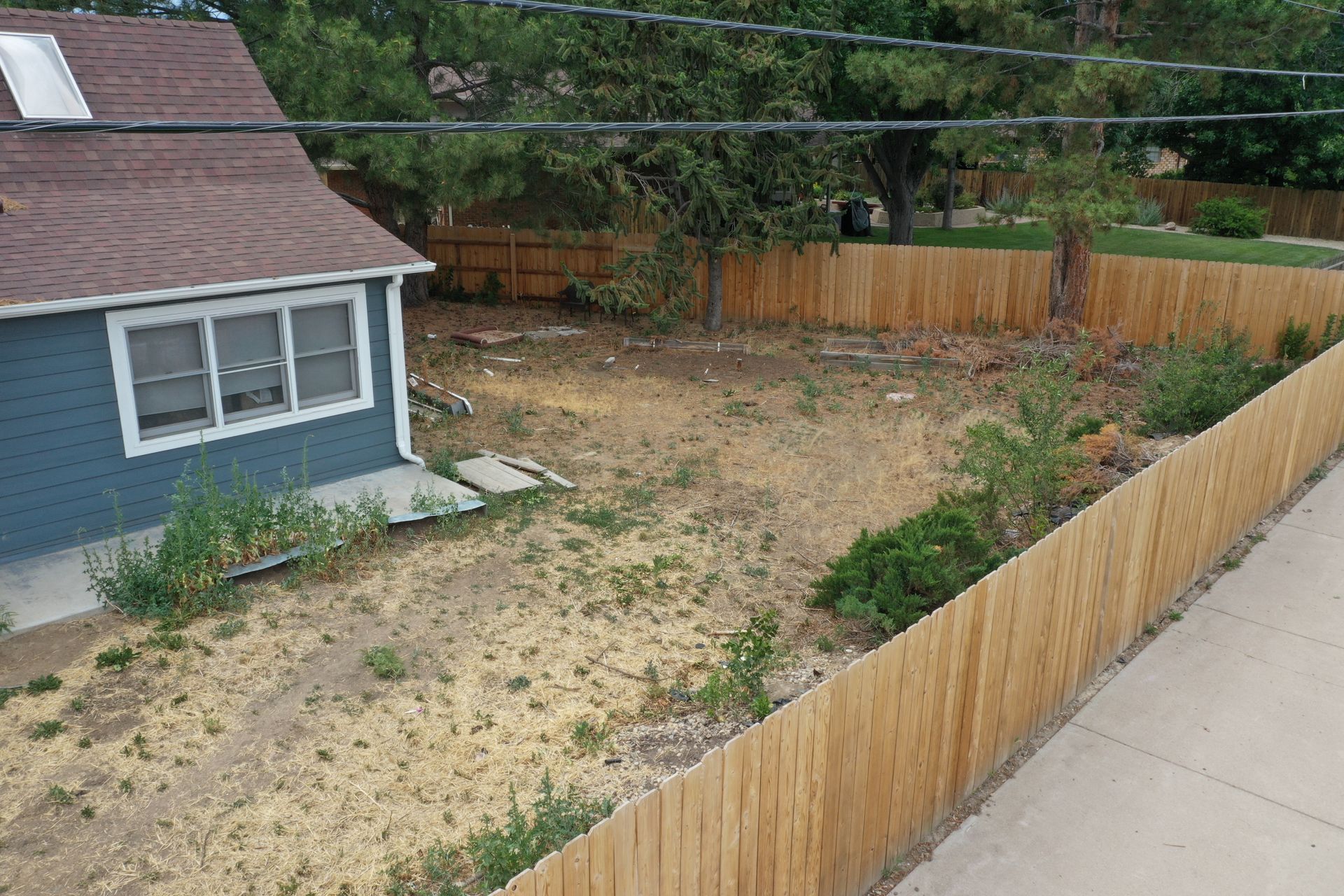 A vacant lot with a wooden fence and a blue house. The ground is dry and patchy with some weeds.