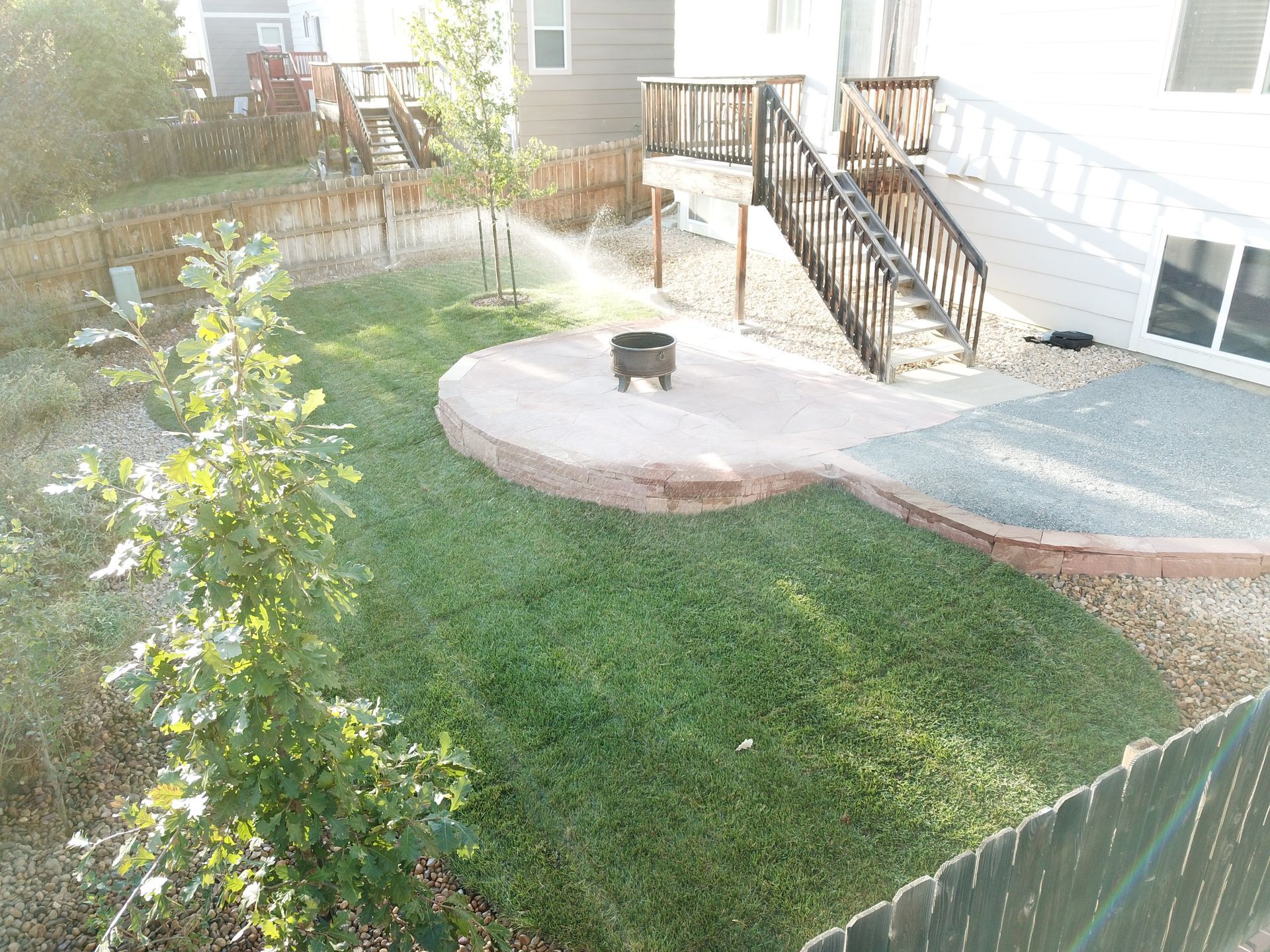 Backyard with a grassy lawn, stone patio, wooden deck, fire pit, and fence.