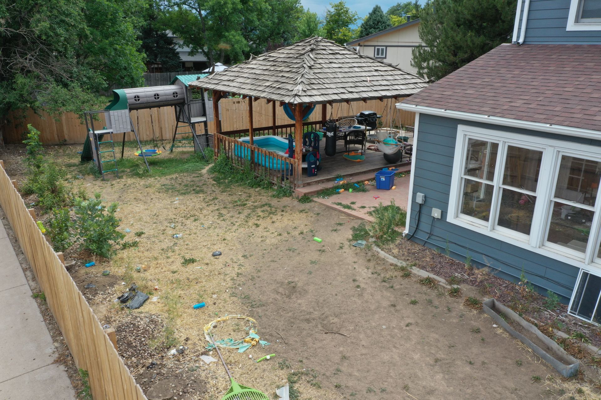 Backyard with gazebo, play set, and blue house. Dry grass and wooden fence.