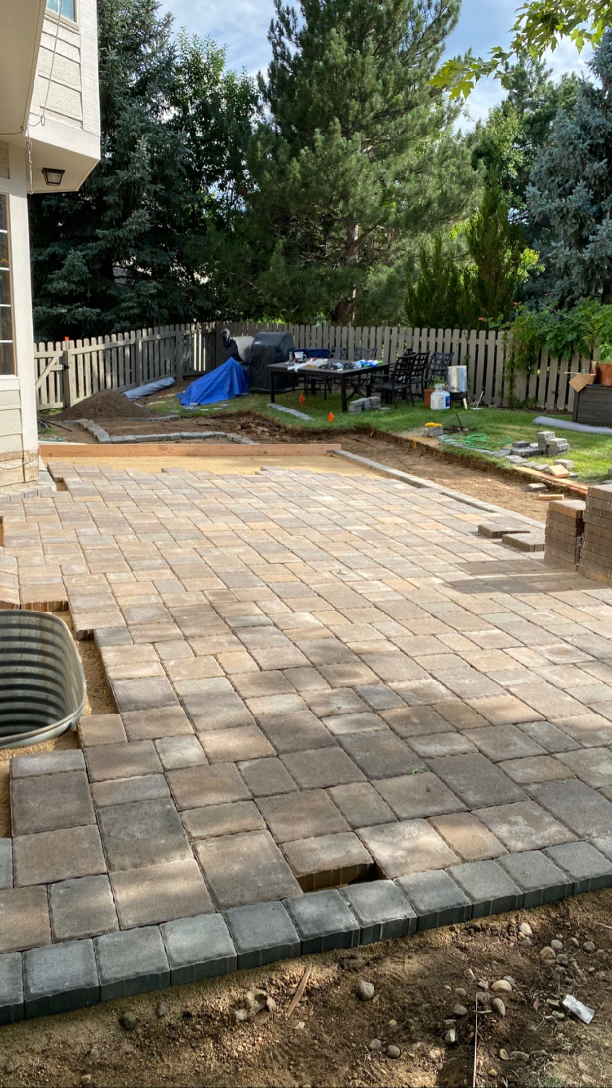 Brick patio under construction, with some bricks laid and others missing. Backyard setting with a fence.