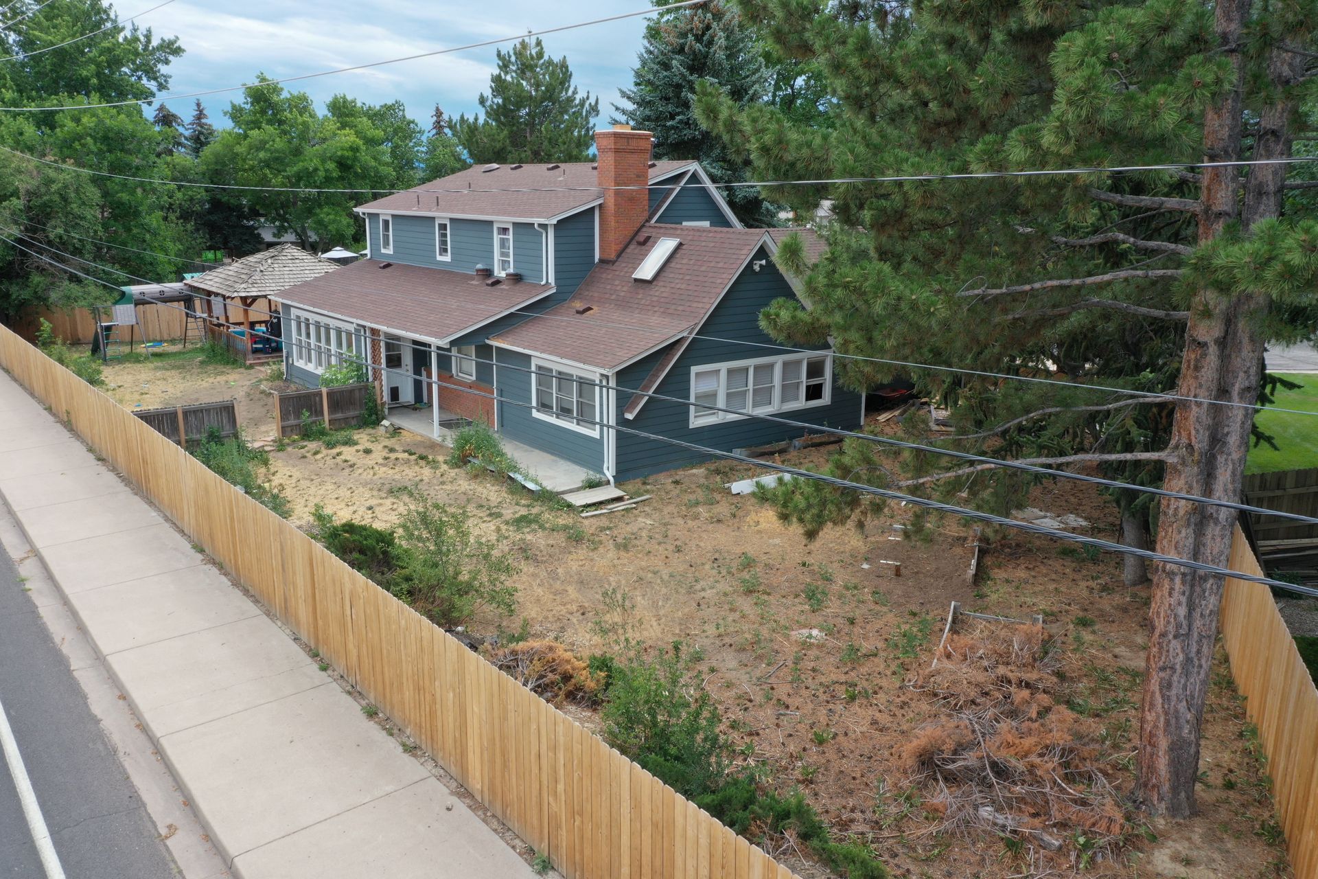 Two-story blue house with brown roof and wooden fence along a road.