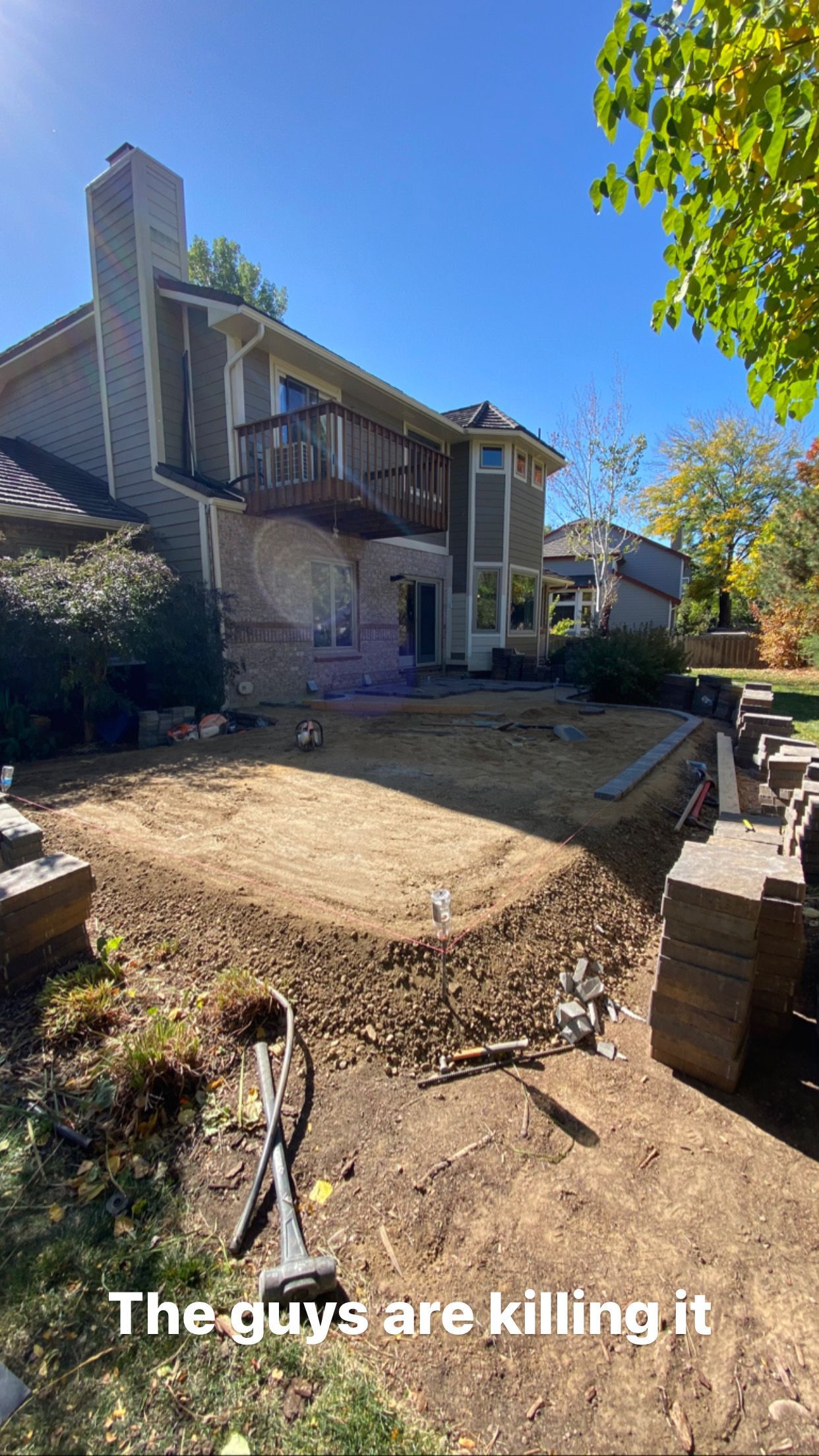 Backyard construction site; dirt, house with deck, tools, and a small machine under a blue sky.