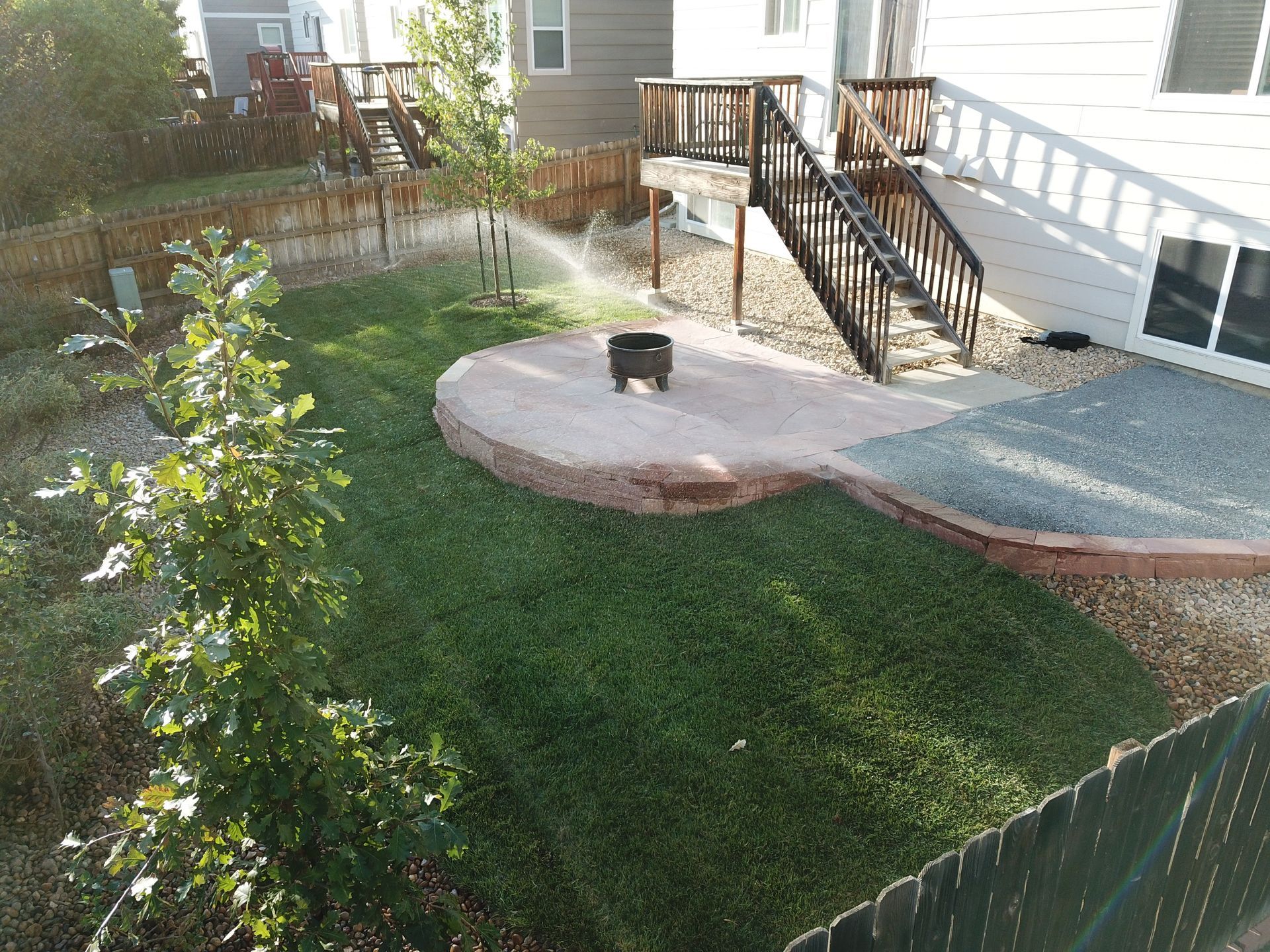 Backyard with green grass, stone patio with fire pit, wooden deck, and a fence.