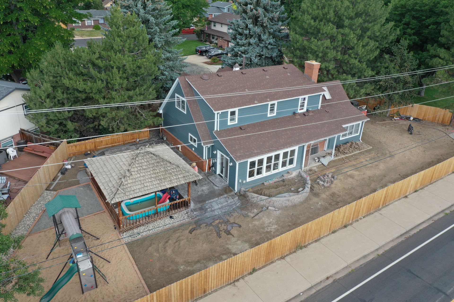 Aerial view of a two-story blue house with a brown roof, surrounded by a wooden fence, with a playground and gazebo.