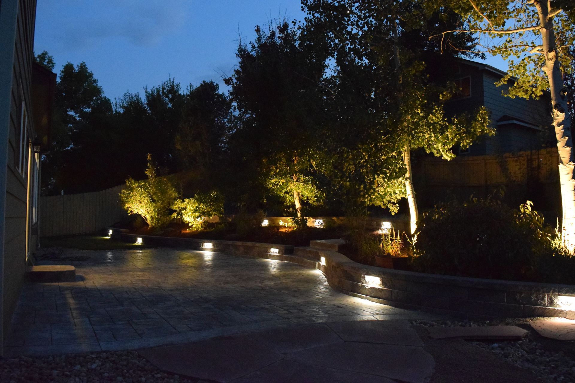 Backyard patio at night, illuminated by soft lights. Trees and landscaping are lit with spotlights.