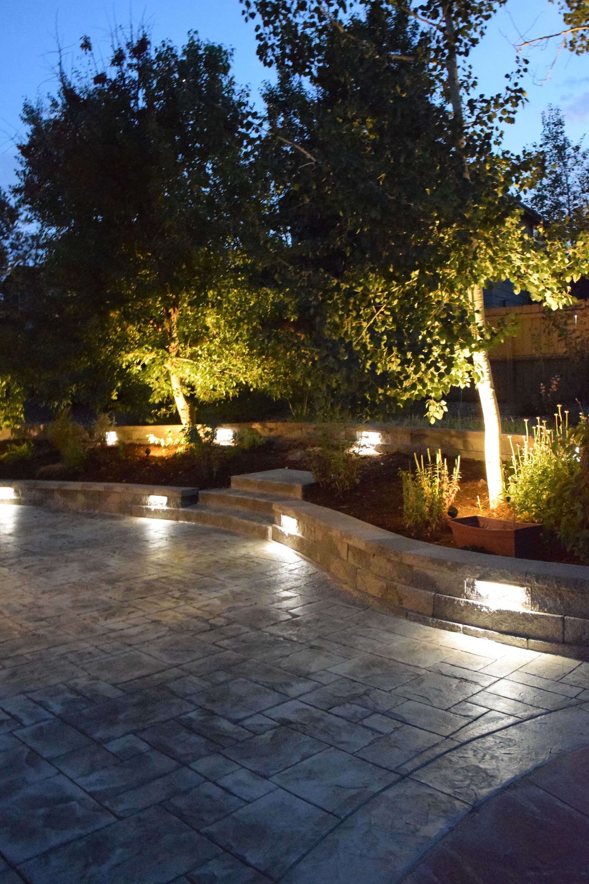 Outdoor patio at dusk, with illuminated trees, retaining walls, and pathway lights.
