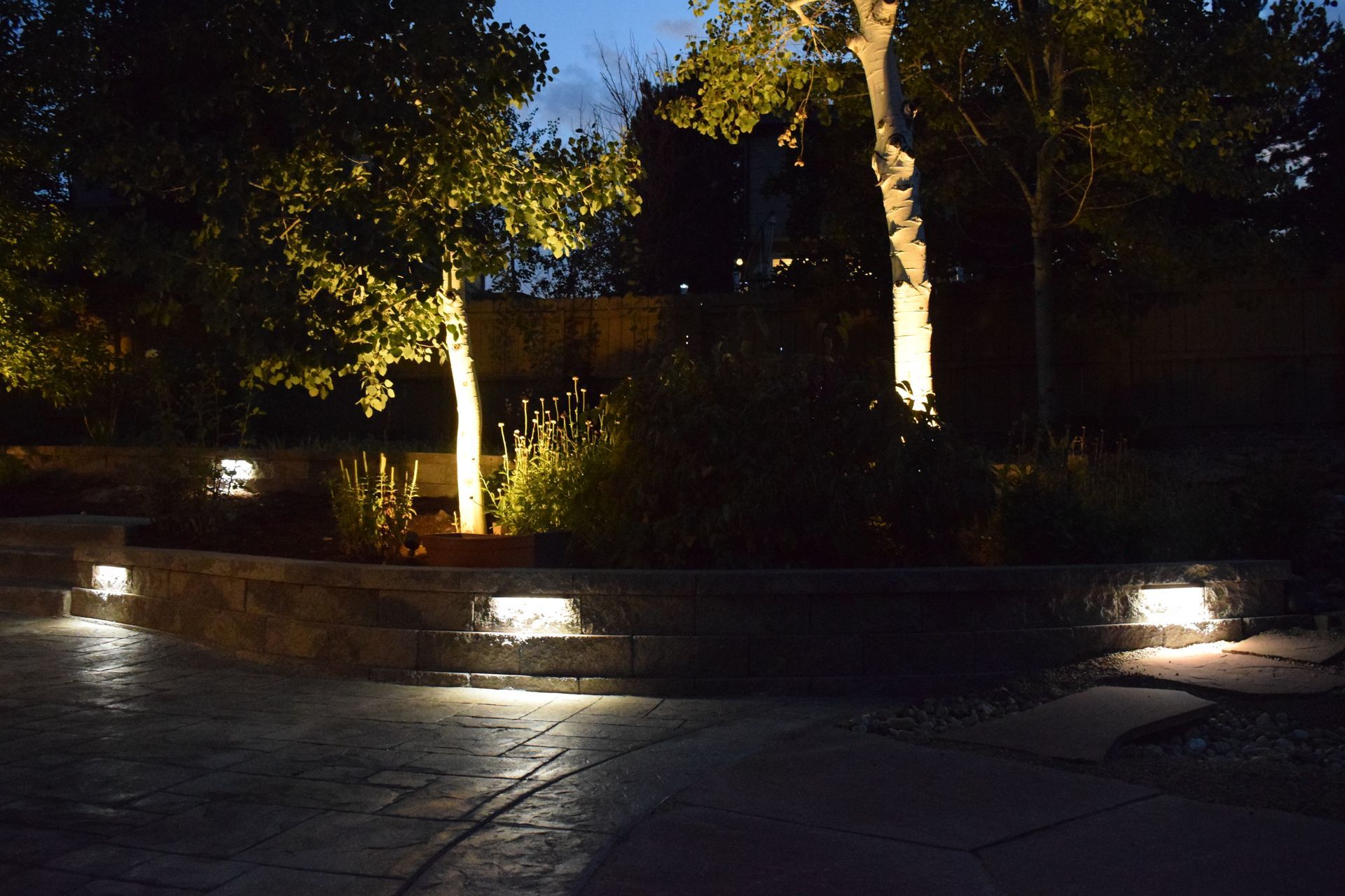 Lit outdoor landscaping at dusk: trees and retaining wall with recessed lights illuminate a patio.