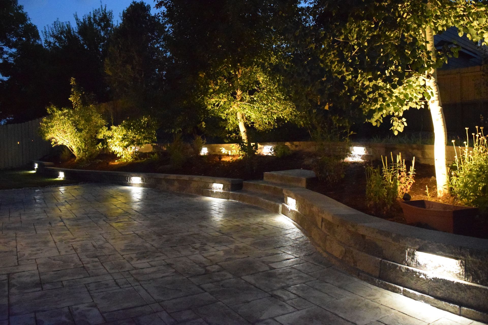 Illuminated backyard at dusk with path lights, stone wall, and trees.