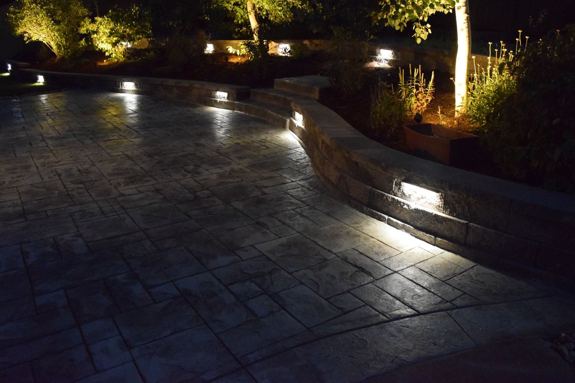 Nighttime view of lit outdoor patio with stone retaining wall. Several lights illuminate the ground and surrounding trees.