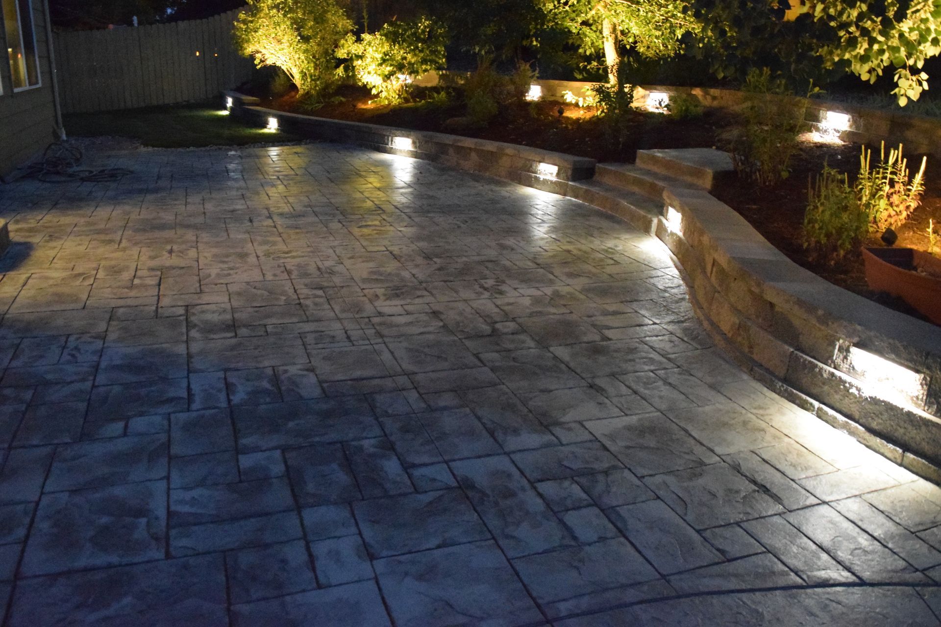 Lit concrete patio with built-in lights, curving stone wall, and illuminated landscaping at night.
