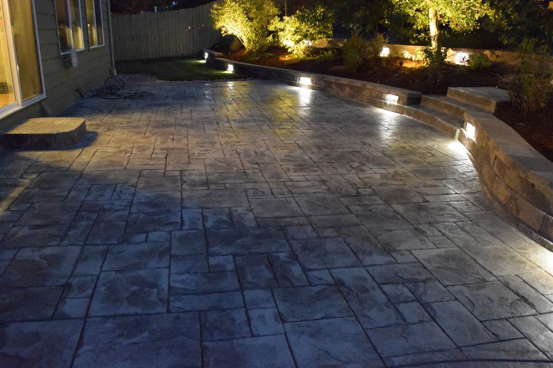 Nighttime view of a stone patio with built-in lights, retaining wall, and backyard landscaping.