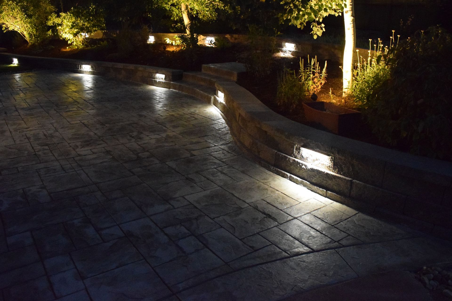 Nighttime view of a patio with stone wall lit by small rectangular lights. Trees in background are also illuminated.