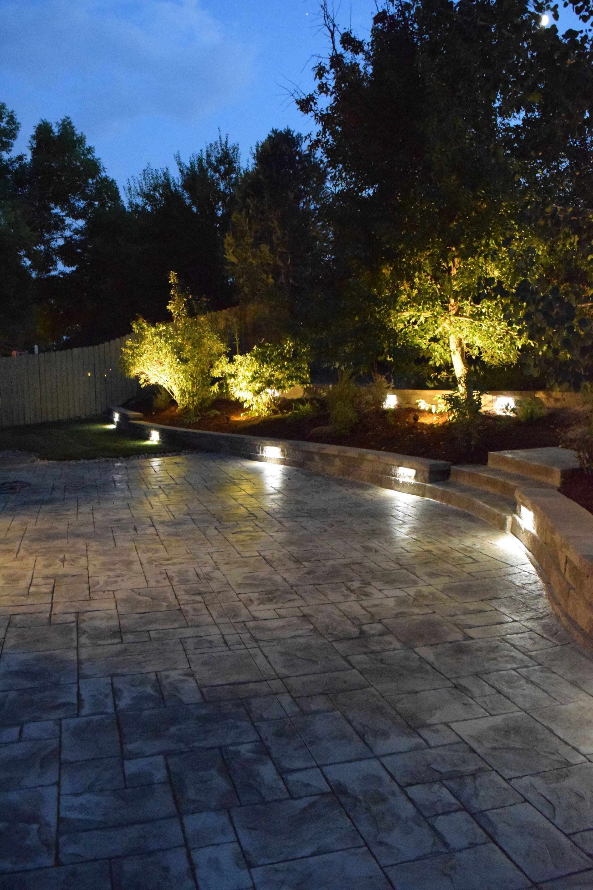 Lit patio at dusk with trees and landscape lighting.