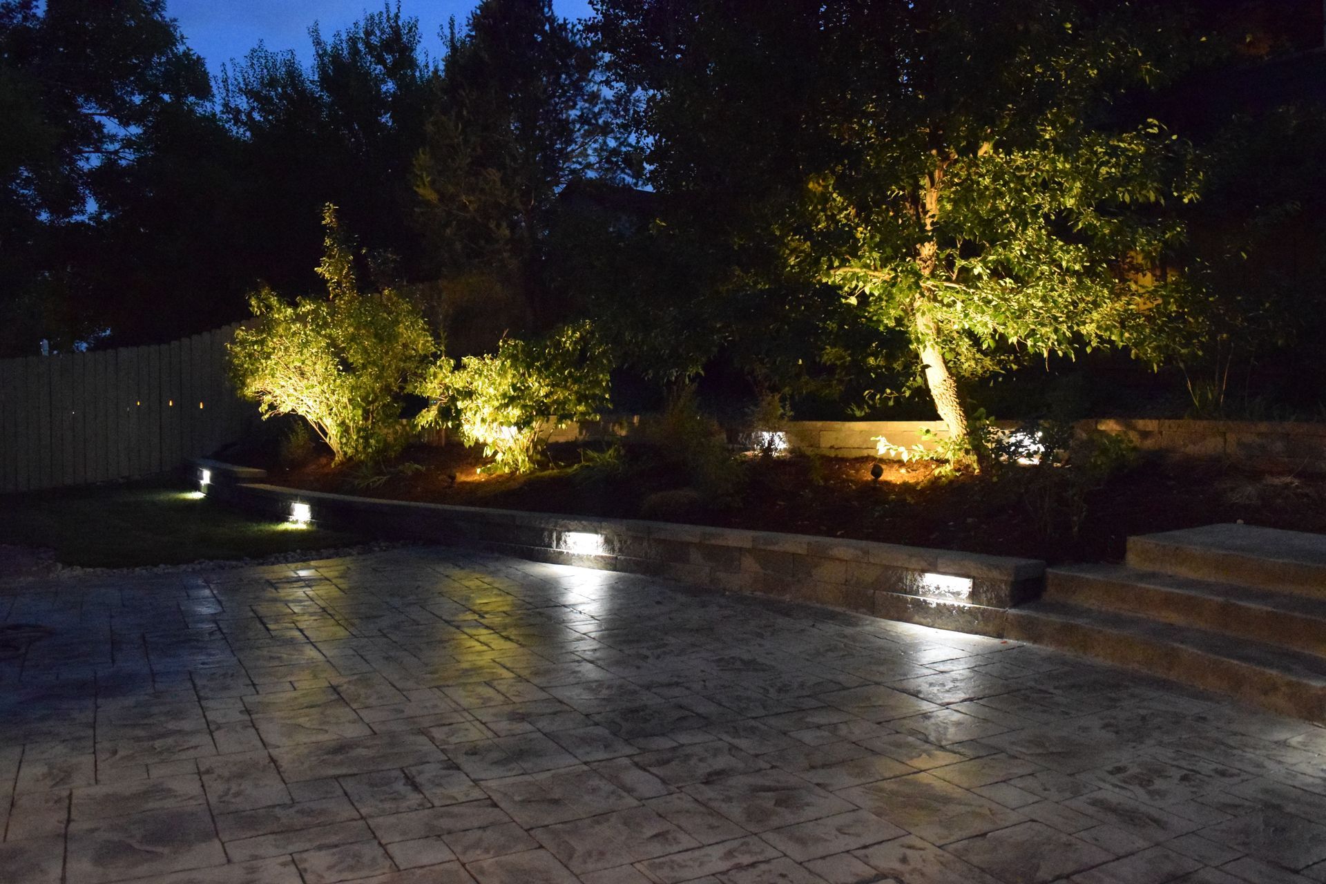 Nighttime landscape with outdoor lighting illuminating trees, bushes, and a stone patio.