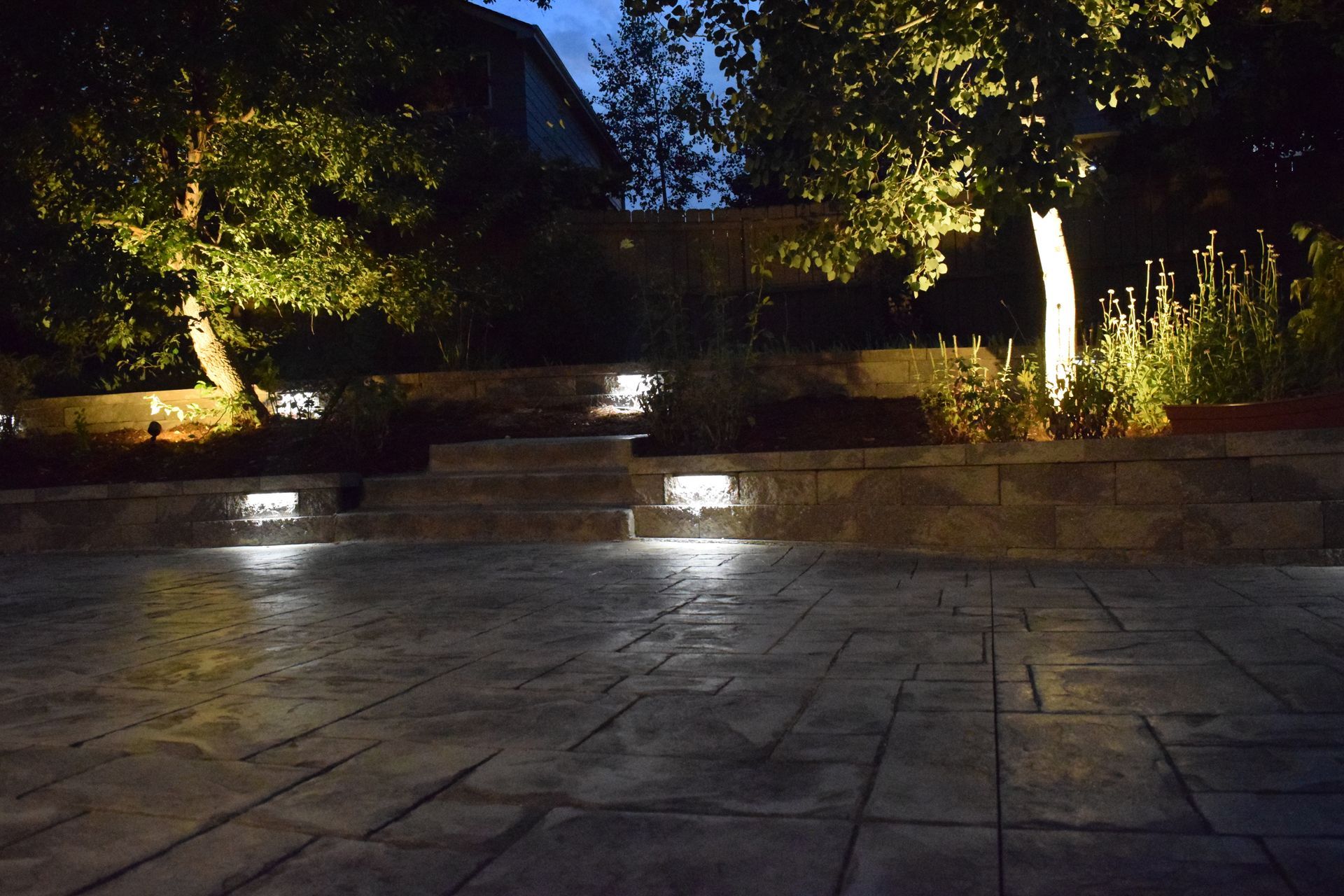 Nighttime view of a patio with built-in lights, tiered retaining wall, and illuminated trees.